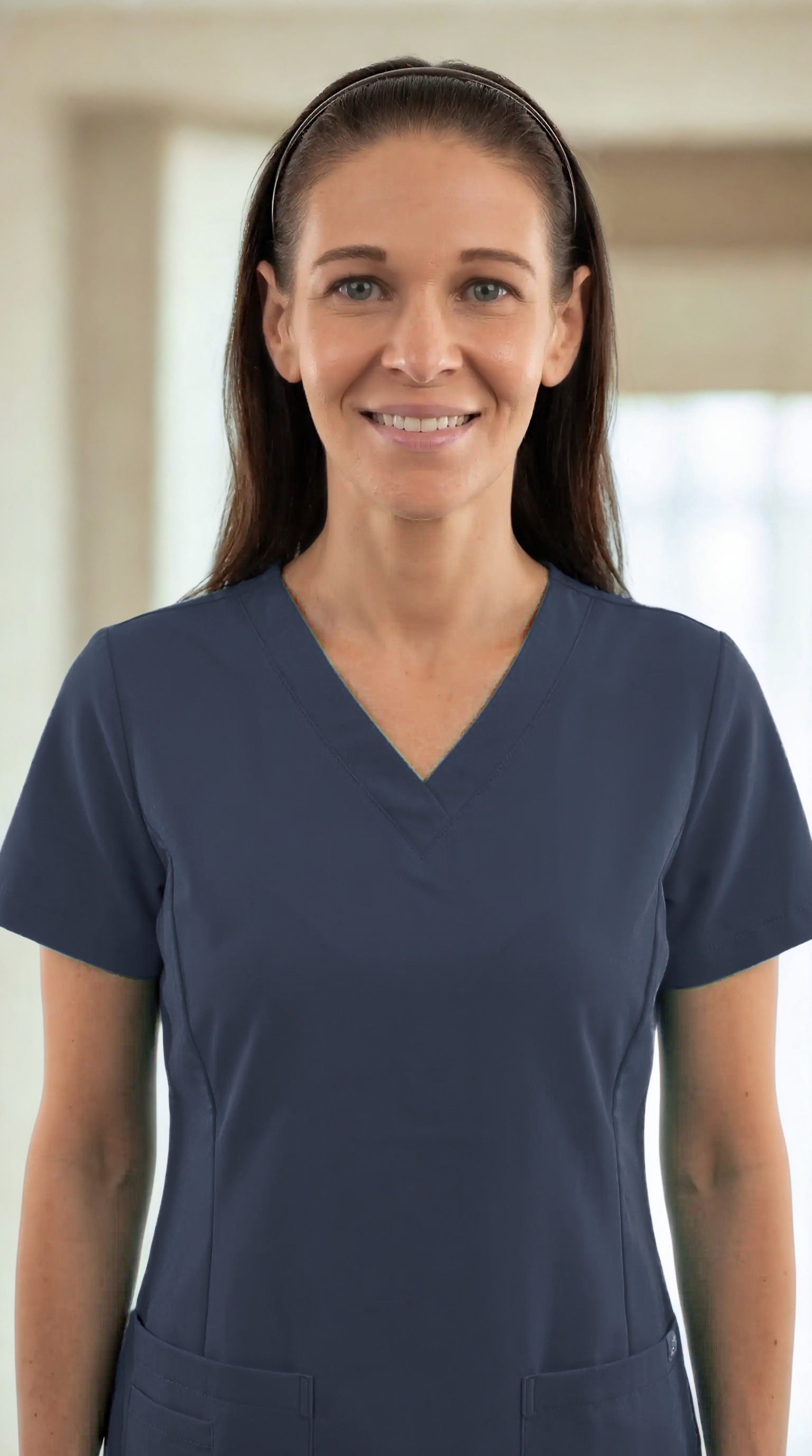 A woman in navy medical scrubs and a headband smiling, standing indoors with a blurred background of windows and curtains.