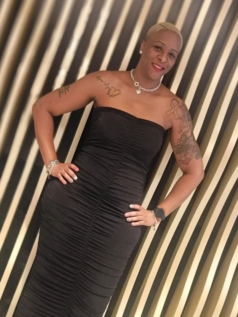 Woman with short platinum blonde hair wearing a strapless black dress, standing in front of a striped background, smiling, accessorized with jewelry, tattoos on arms, and a smartwatch.