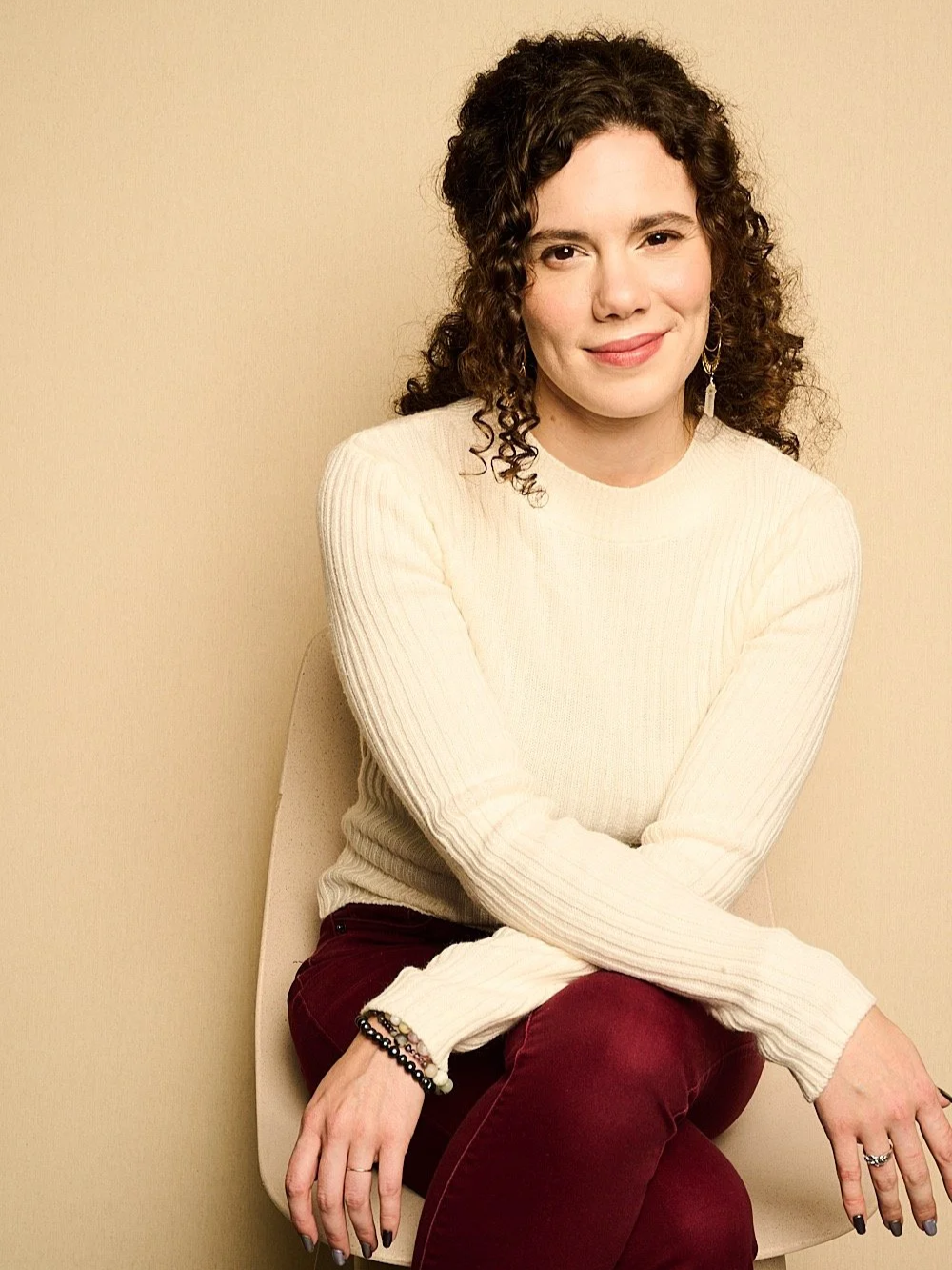 A woman with curly brown hair sitting on a beige chair against a beige background, wearing a cream sweater and burgundy pants, smiling slightly at the camera.