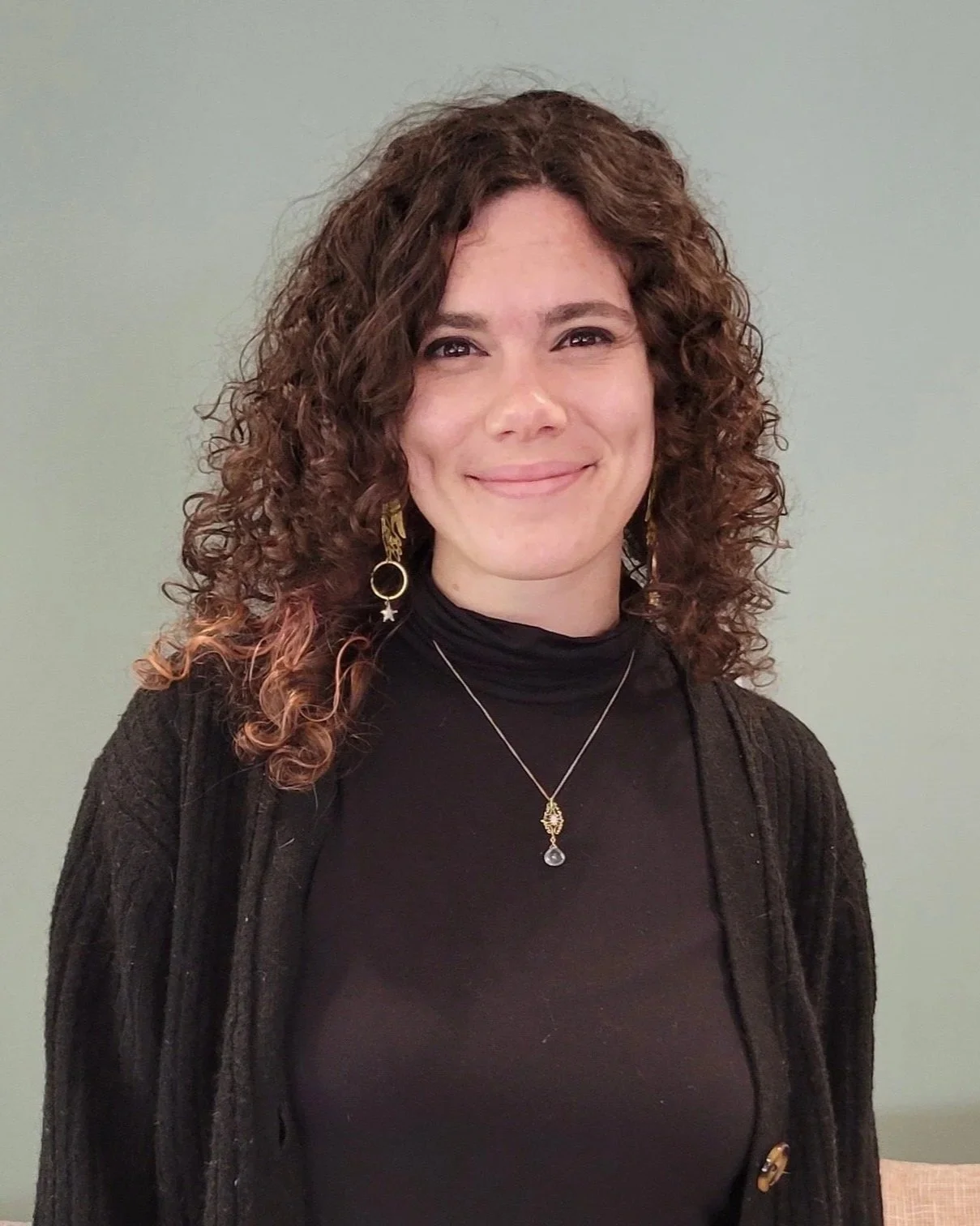 A therapist with shoulder-length curly brown hair, wearing a black turtleneck and a black cardigan, smiling at the camera. She has gold earrings and a necklace with a gold and aquamarine pendant.