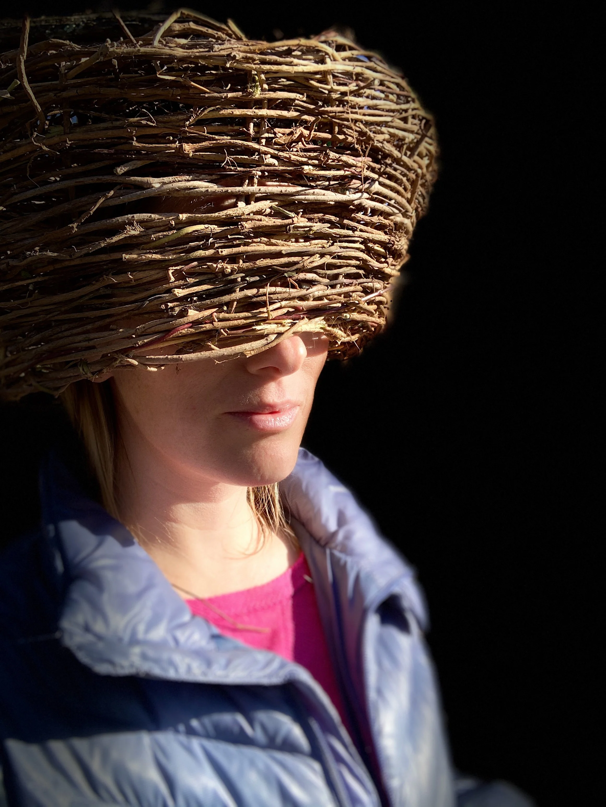 Person wearing a large woven basket on head, partially obscuring face, with pink top and light jacket, against dark background.