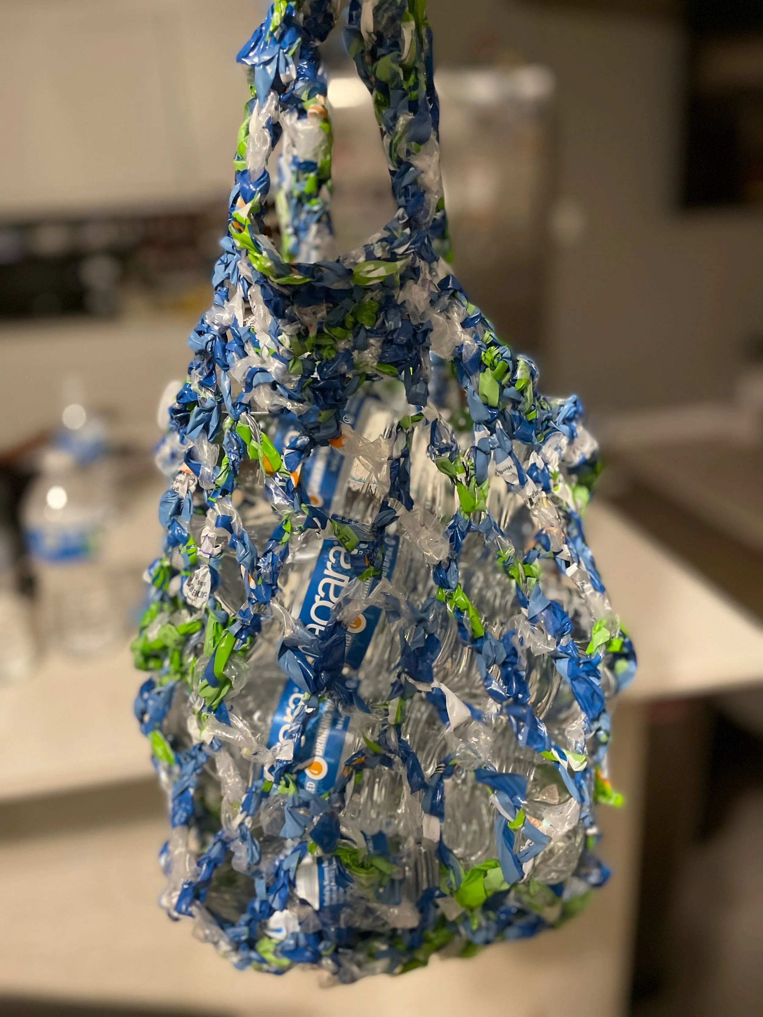 A handmade bottle holder made from blue, green, and white plastic twist ties, designed to hold a bottle of sparkling water.