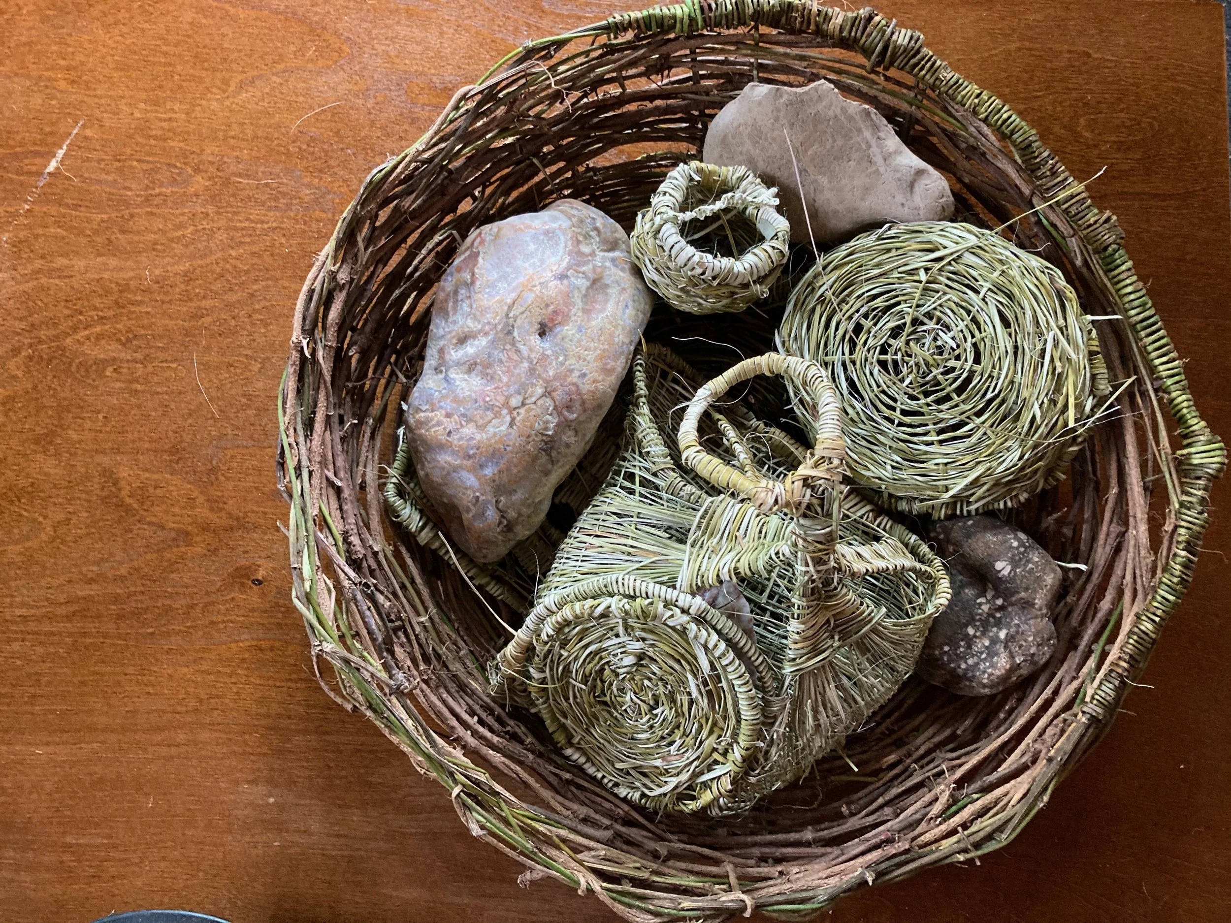 A woven basket containing rocks, seashells, and small decorative woven items on a wooden surface.