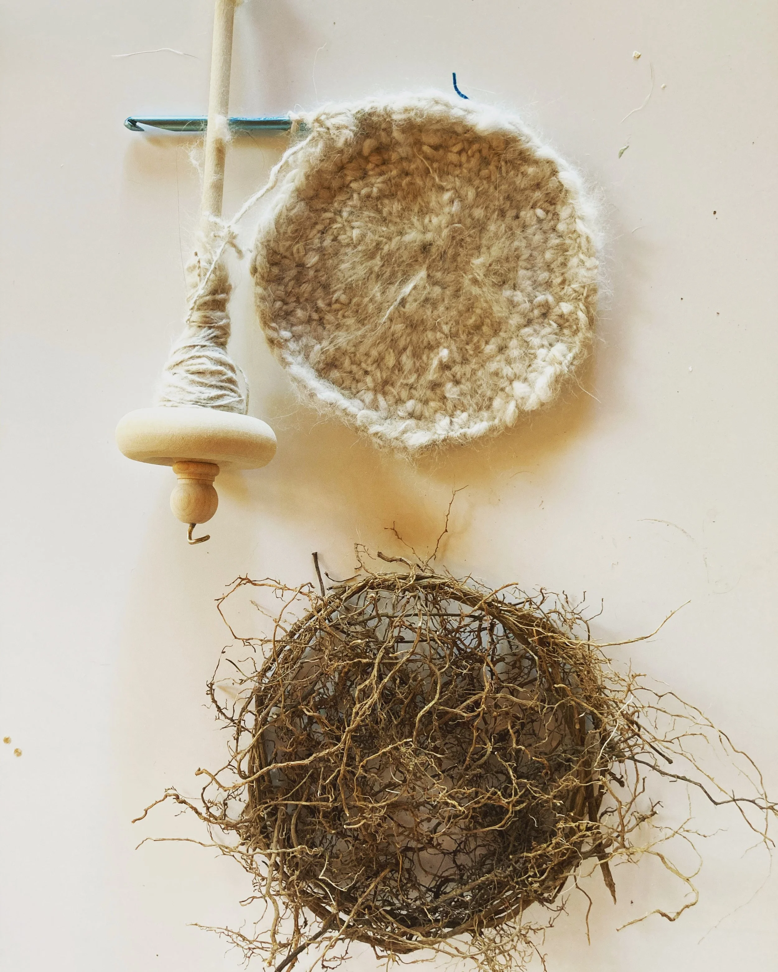 A woodworking spinning wheel, a wool yarn bowl, and a wreath of dried twigs arranged on a light-colored surface.