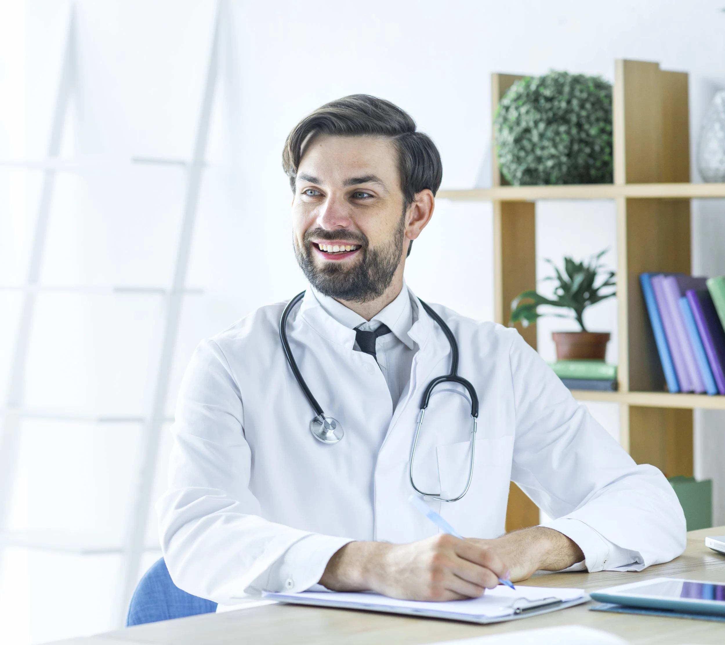 A male doctor with a stethoscope around his neck sitting at a desk with a notebook and tablet, smiling in a bright medical office with shelves of books and plants in the background.