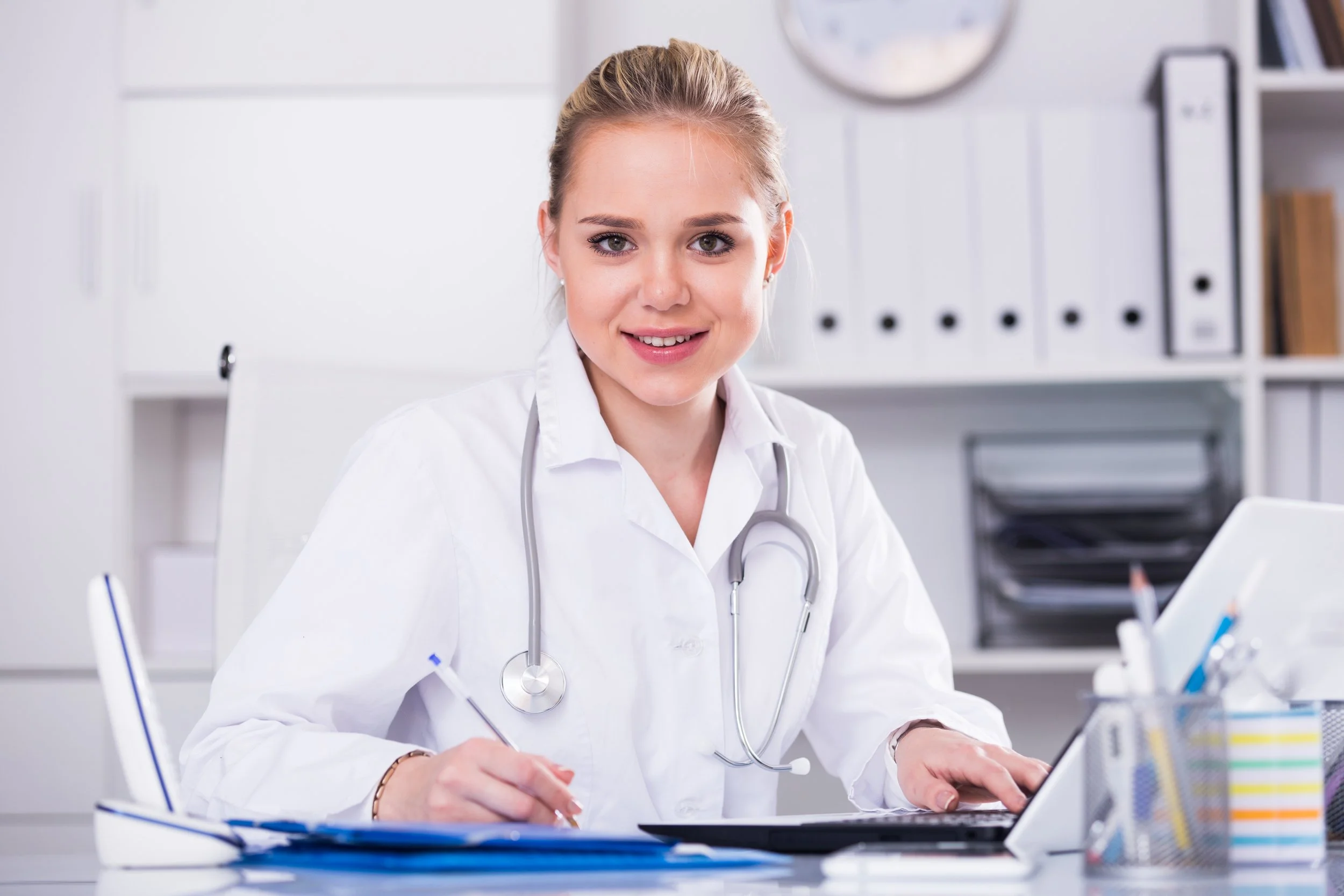 A young female doctor sitting at an office desk, wearing a white coat and stethoscope, smiling at the camera while holding a pen and working on a tablet.