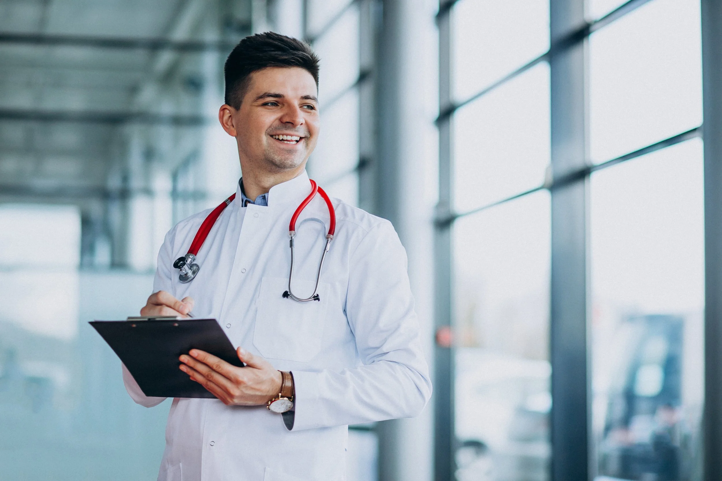 A smiling male doctor or healthcare professional in a white coat with a red stethoscope around his neck, holding a clipboard, standing in a modern building with large windows.
