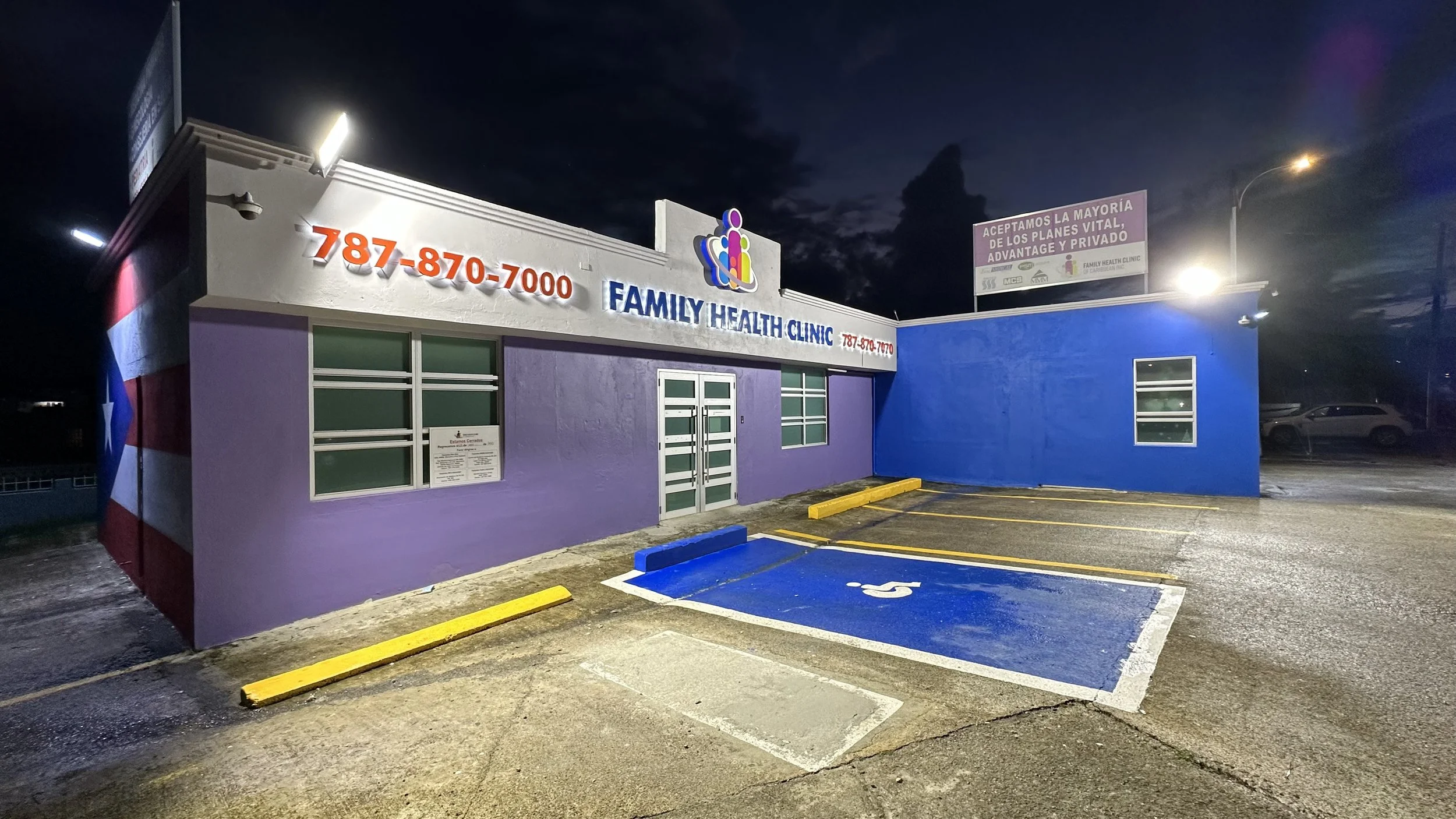 Parking lot outside a Family Health Clinic at night with a blue handicapped parking space and a purple and white building with yellow curbs.