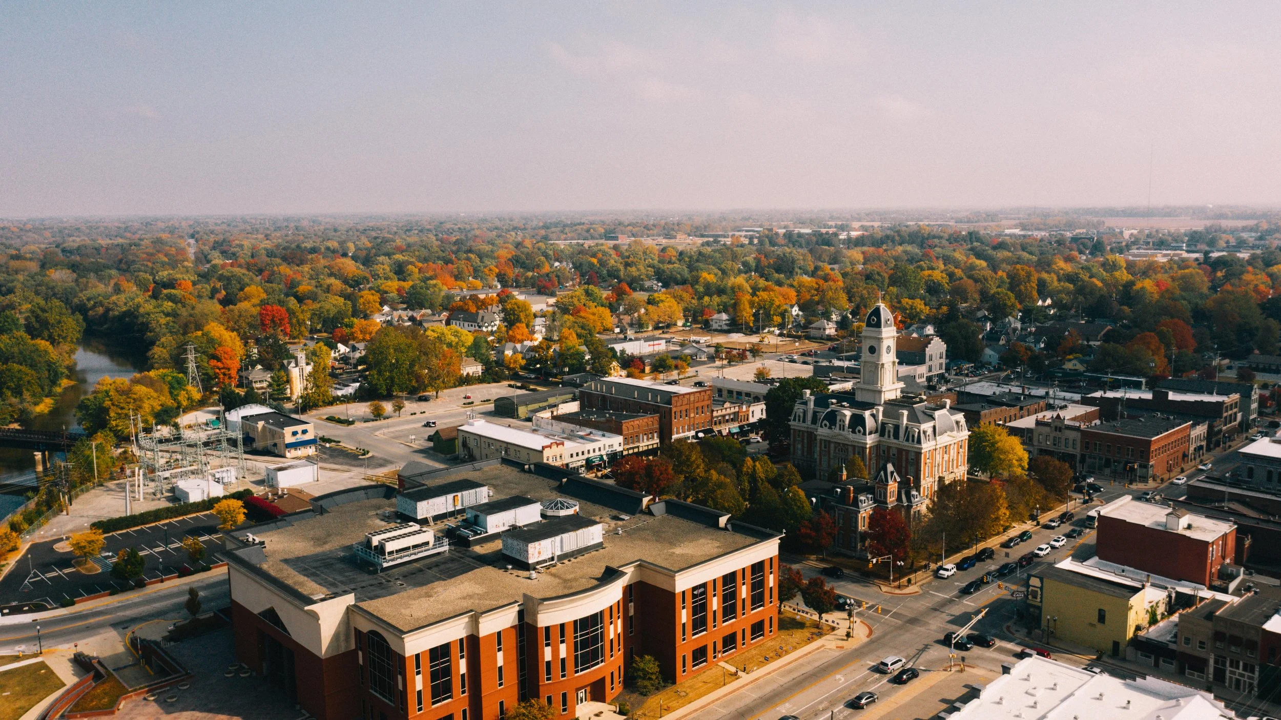 Aerial view of a small downtown area with historic buildings, a clock tower, and a river, surrounded by colorful fall trees.