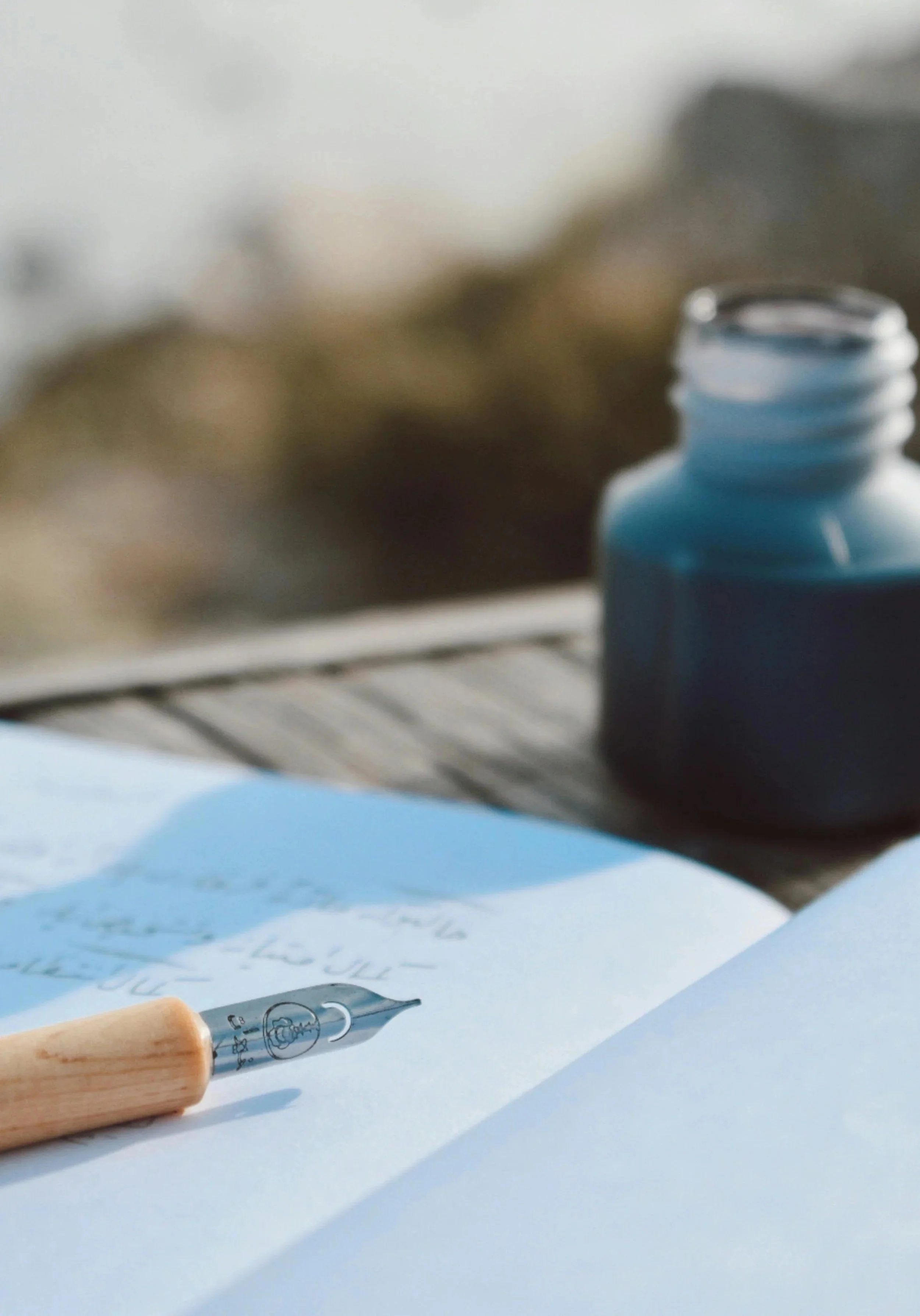 Close-up of a fountain pen with a wooden handle writing on paper with handwritten text, and a blue ink bottle in the background.