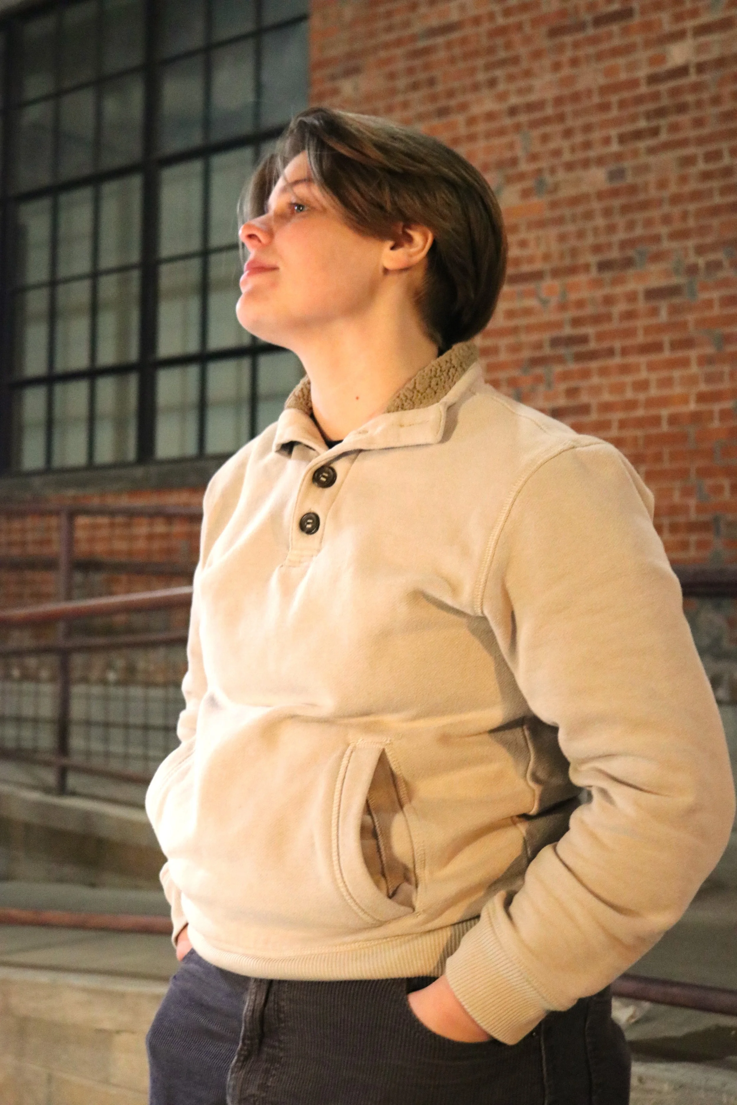 Person with short brown hair wearing a beige sweatshirt with a collar and a front pocket, standing indoors with brick wall and large window in background.