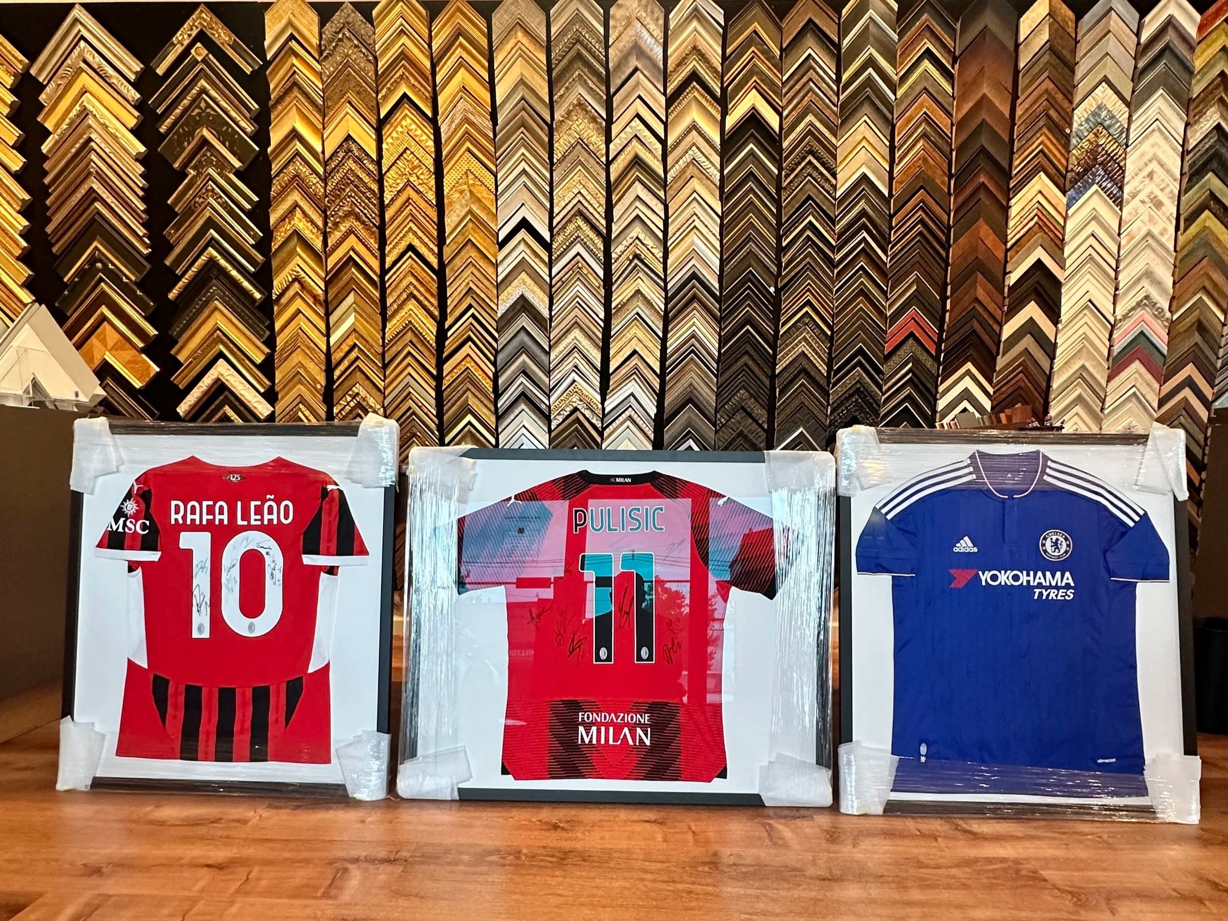 Three framed football jerseys displayed on a wooden surface with a background of various picture frame moldings. The left jersey is red and black, labeled "RAFA LEÃO" with the number 10. The middle jersey is red with black accents, labeled "PULISIC" with the number 11, and includes the word "MILAN" at the bottom. The right jersey is blue with white accents, marked with the Chelsea FC logo and "YOKOHAMA TYRES."