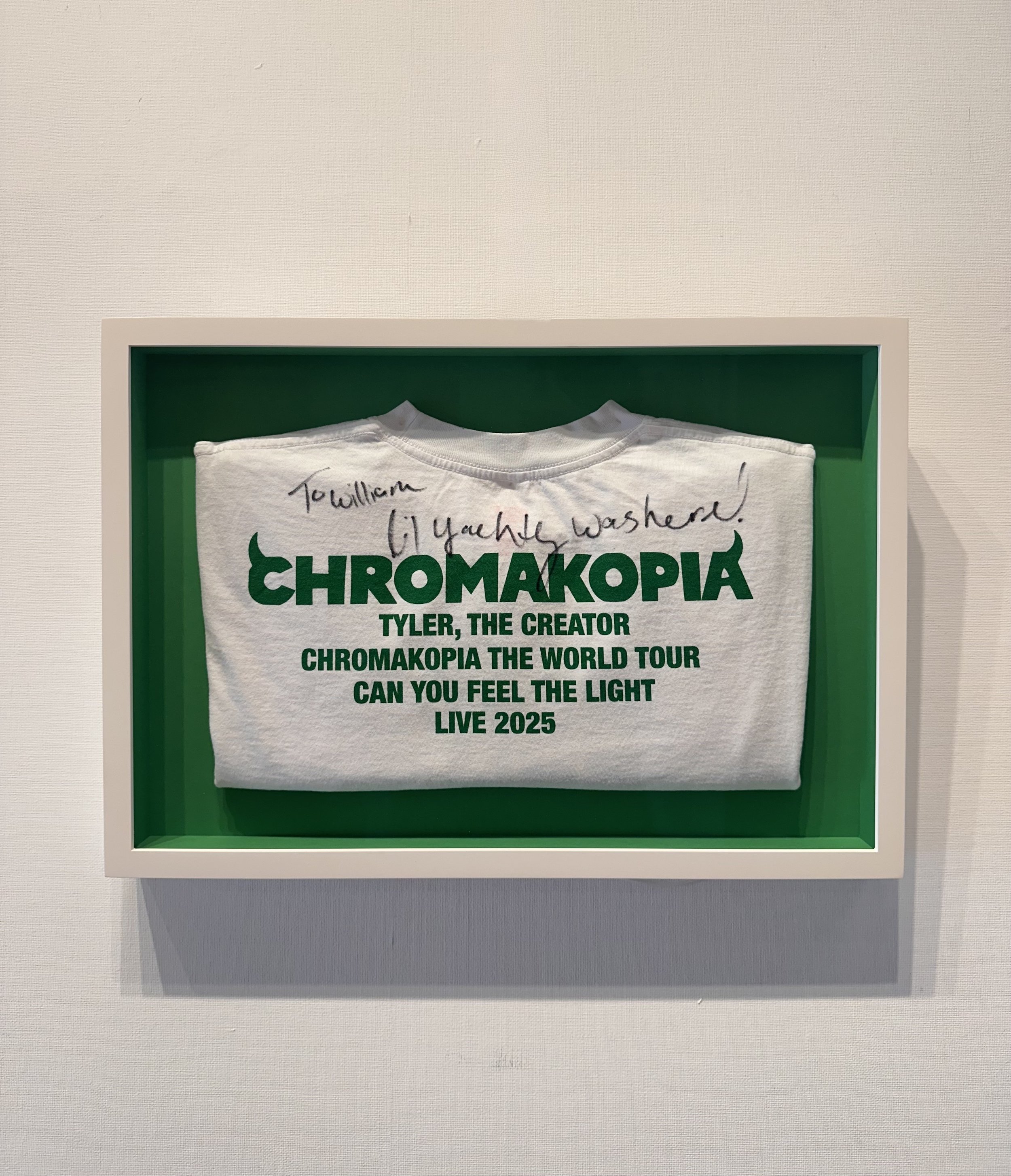 A white T-shirt with green text inside a white shadow box. The shirt has handwritten writing at the top. The printed text reads: 'CHROMAKOPIA TYLER, THE CREATOR CHROMAKOPIA THE WORLD TOUR CAN YOU FEEL THE LIGHT LIVE 2025'.