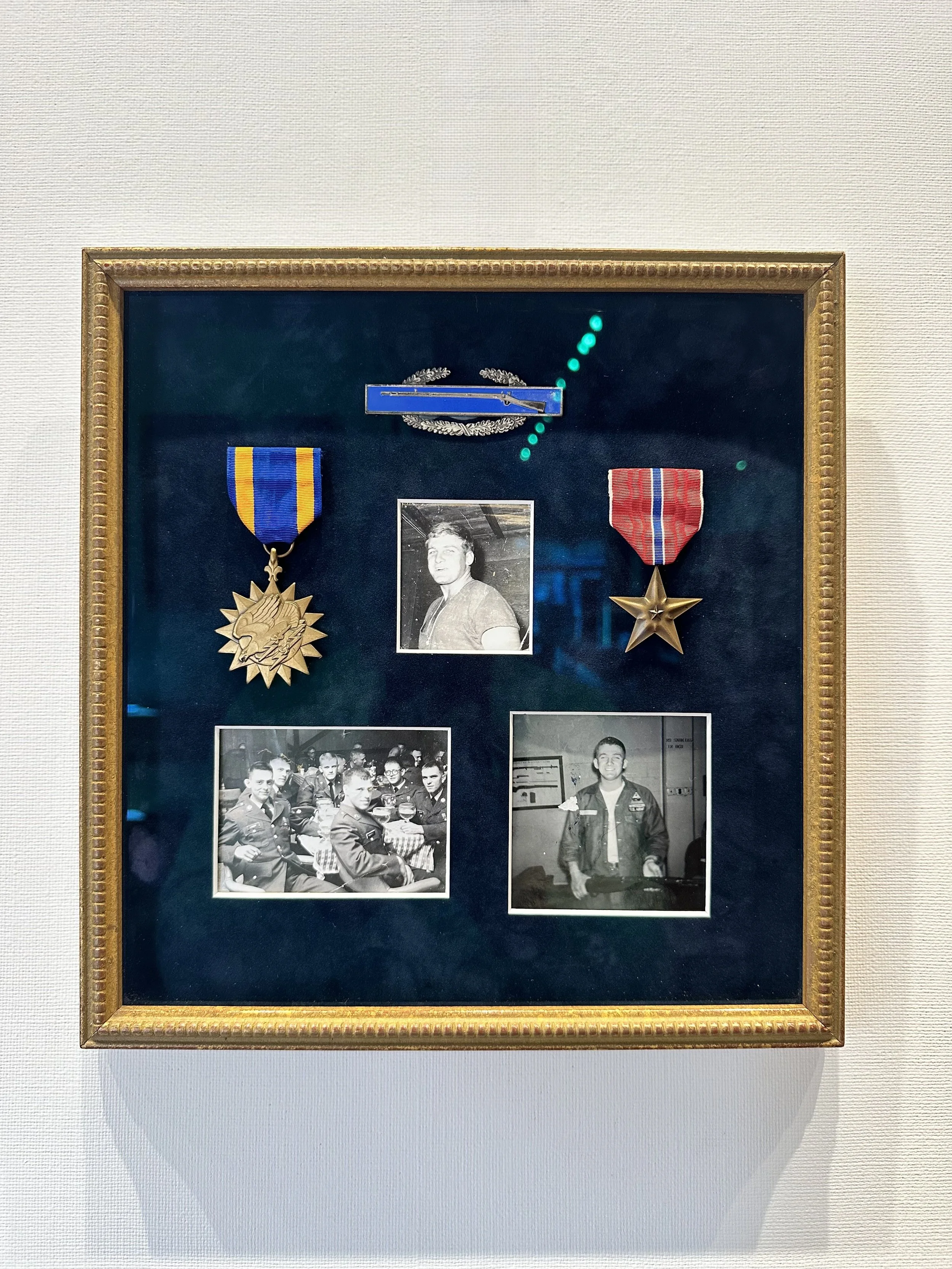 A framed display with two medals, a paper clip, a photograph of a young man, and two black-and-white photographs of groups of young men in military uniforms. The display is mounted on a light-colored wall.