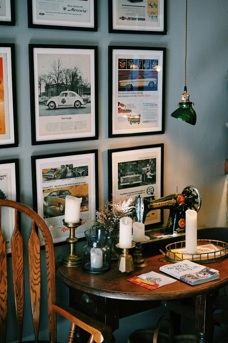 A cozy room corner featuring a wooden table decorated with candles, a vintage sewing machine, and a small bouquet, with framed car advertisements and photos on the wall behind.
