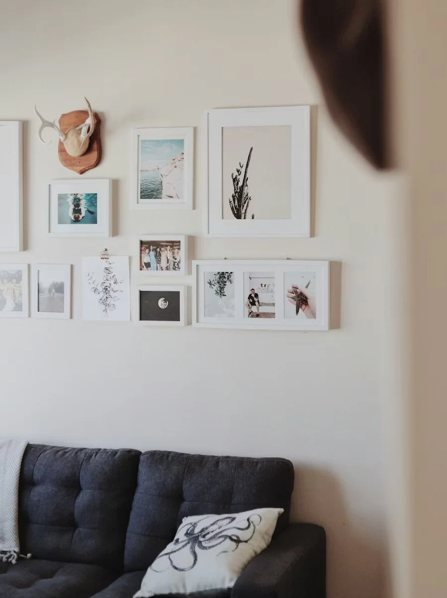 Interior view of a living room featuring a collection of framed photographs and artwork arranged on a white wall above a dark gray couch, with a decorative throw pillow and a partial view of a person's head in the foreground.
