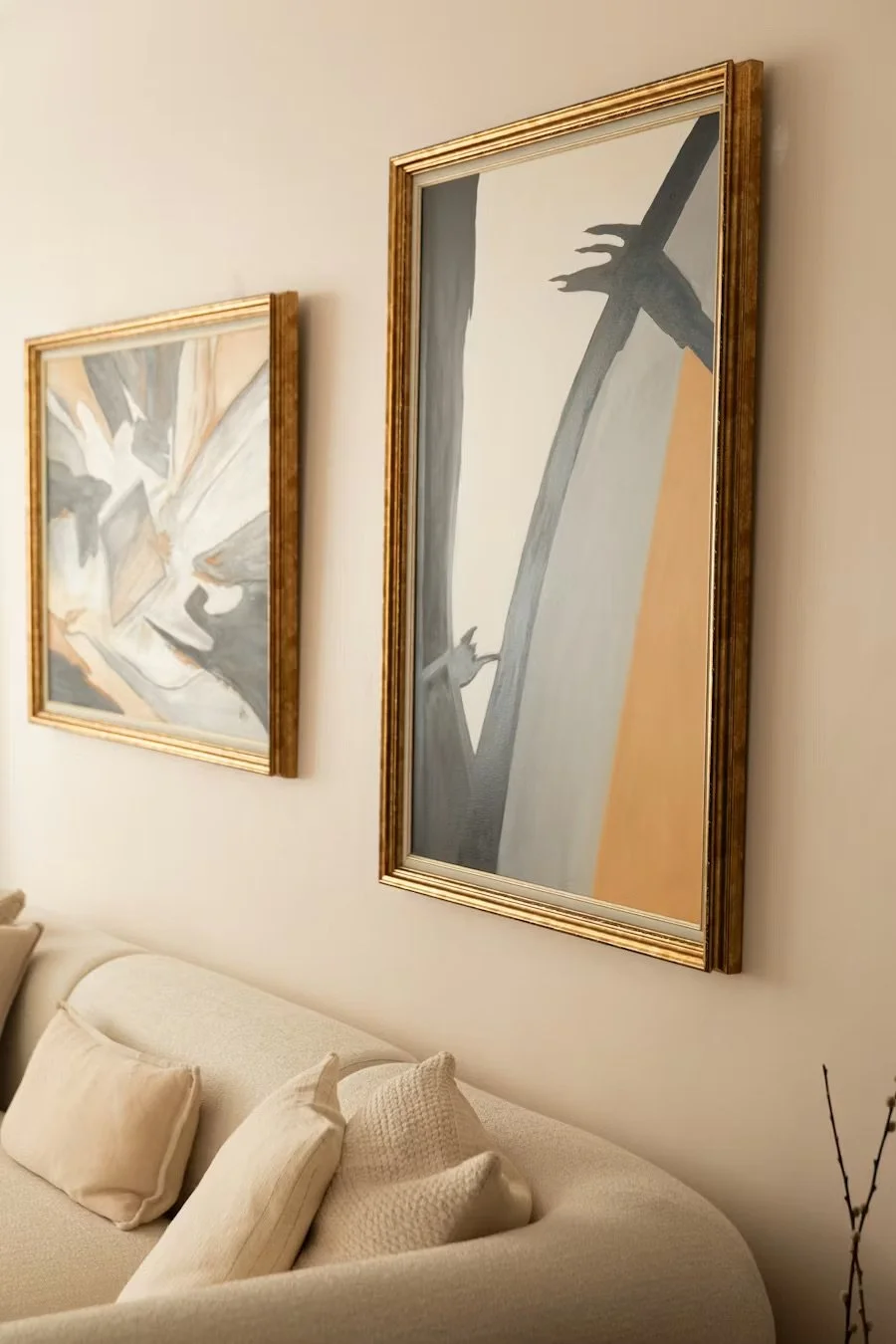 Two abstract paintings in gold frames hanging on a wall above a white sofa with beige pillows.