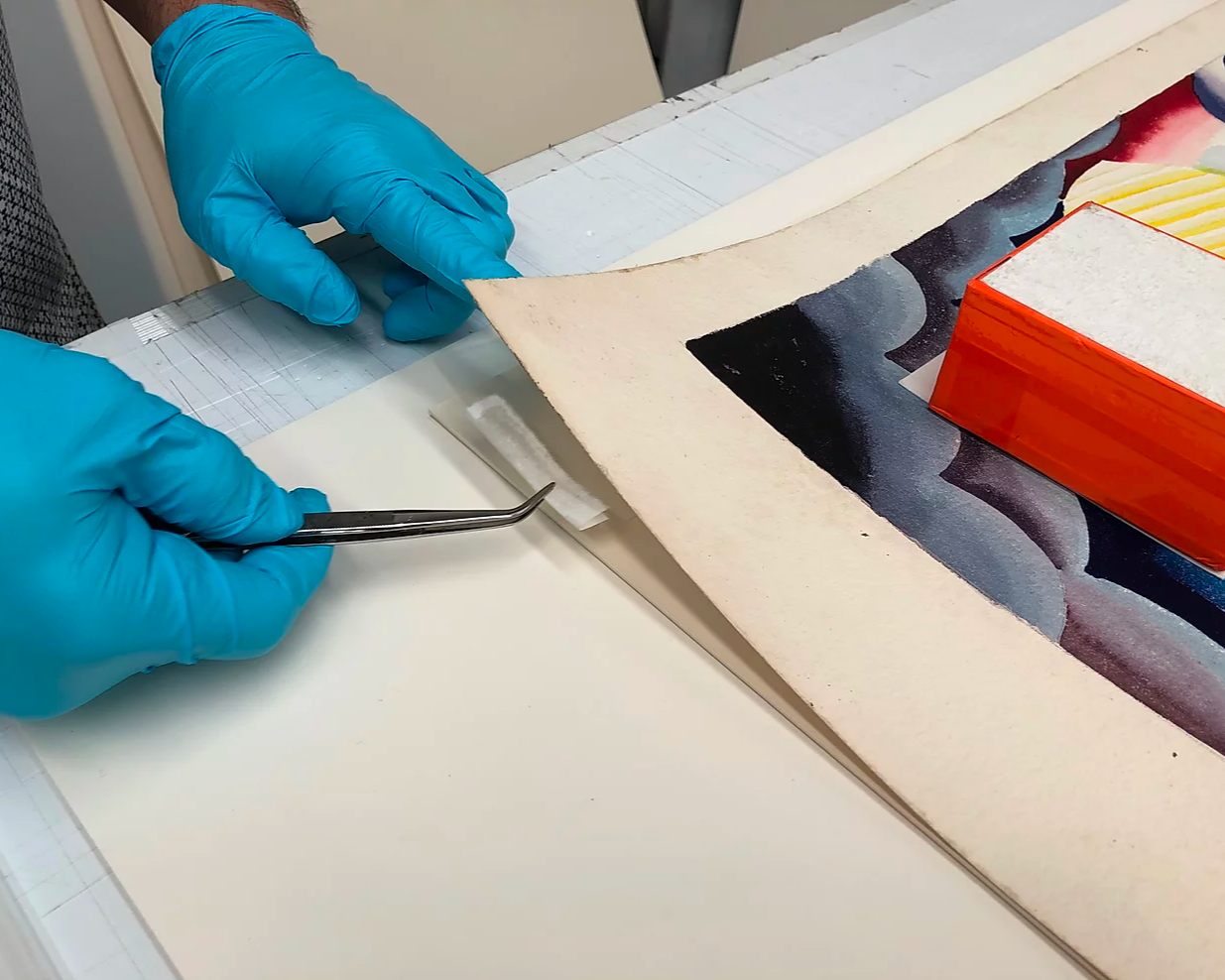 A person wearing blue gloves handling a piece of art on a flat surface with tweezers inside a conservation or framing workspace.
