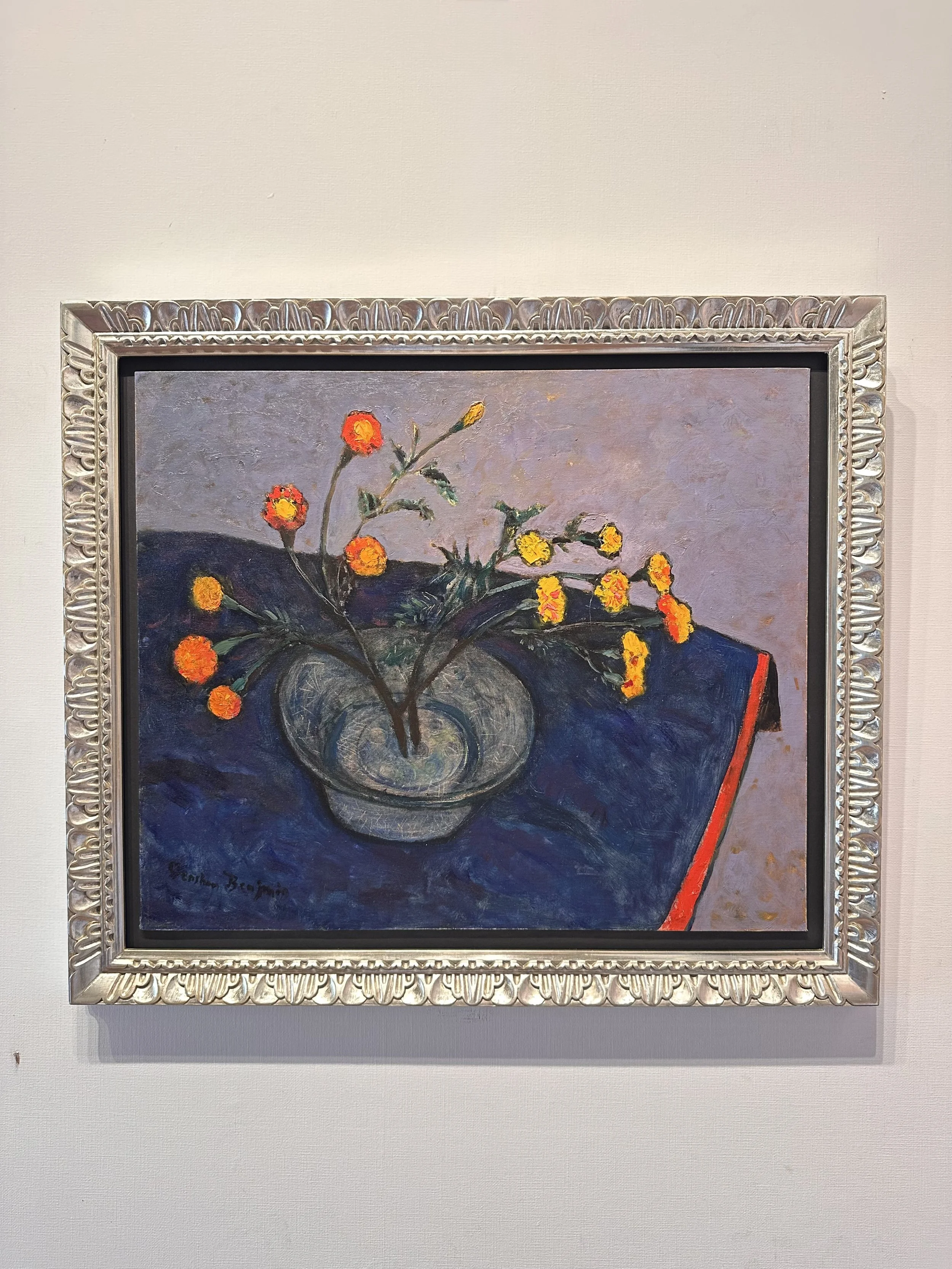 A still life painting of a glass vase with yellow and orange flowers on a dark blue cloth with a light purple background.