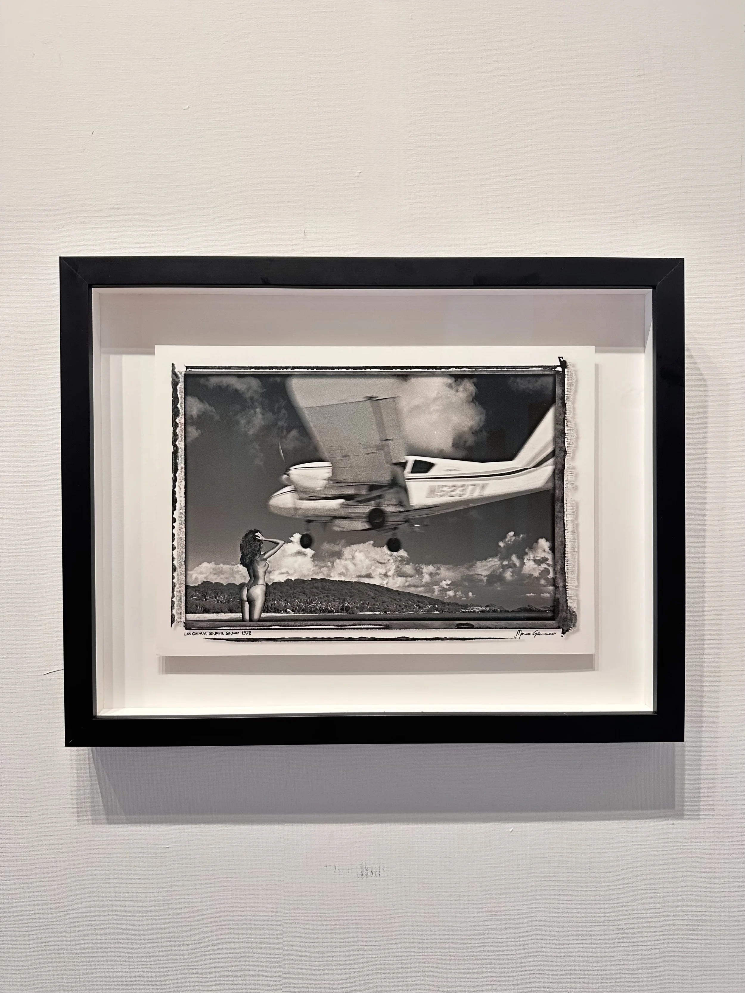 Black and white photograph of a woman standing underneath an airplane flying low, with hills and clouds in the background.