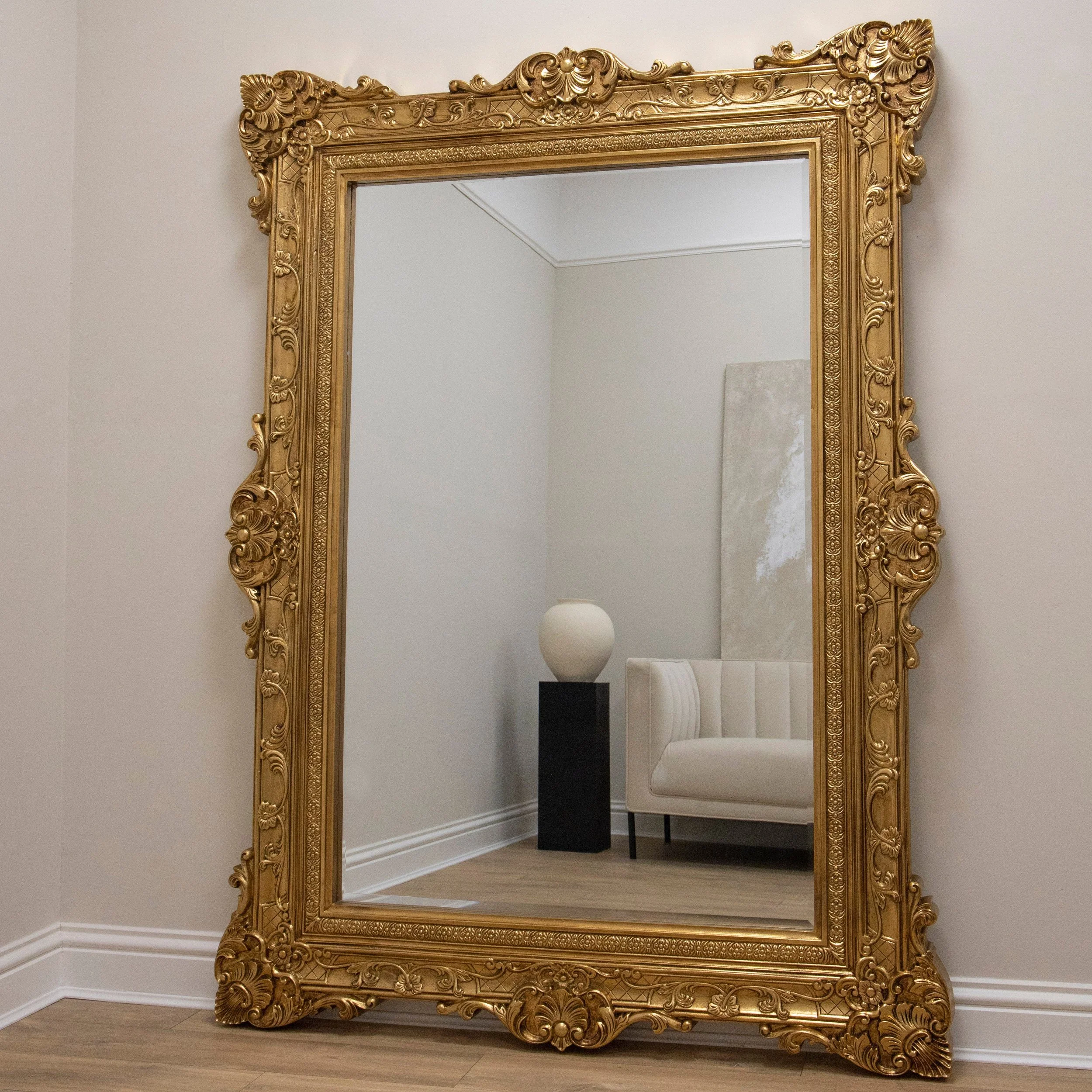 Large ornate gold framed mirror reflecting a white corner with a white armchair, a black pedestal with a white vase, and a rectangular abstract art piece on the wall.