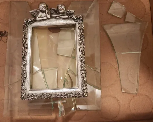 Broken mirror with ornate silver frame and two cherubs at the top, lying on a wooden surface with broken glass pieces nearby.