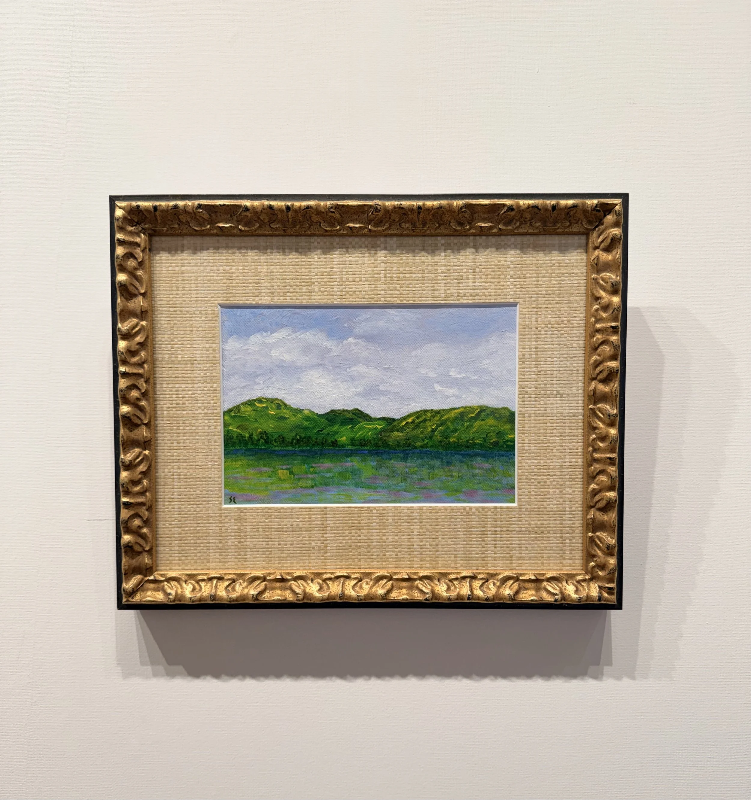 Small landscape painting of green hills and a lake, with a partly cloudy sky, framed in an ornate gold frame, hung on a white wall.