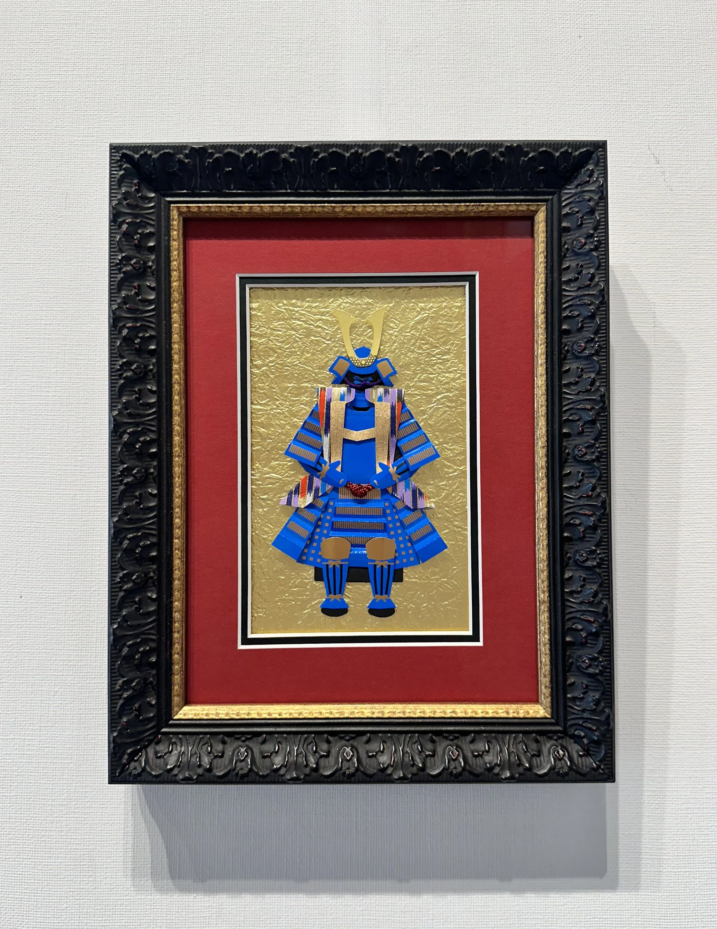 Framed artwork depicting a stylized Anubis figure in bright colors with gold, blue, and red details on a gold textured background, surrounded by a decorative black and gold frame and a red mat.