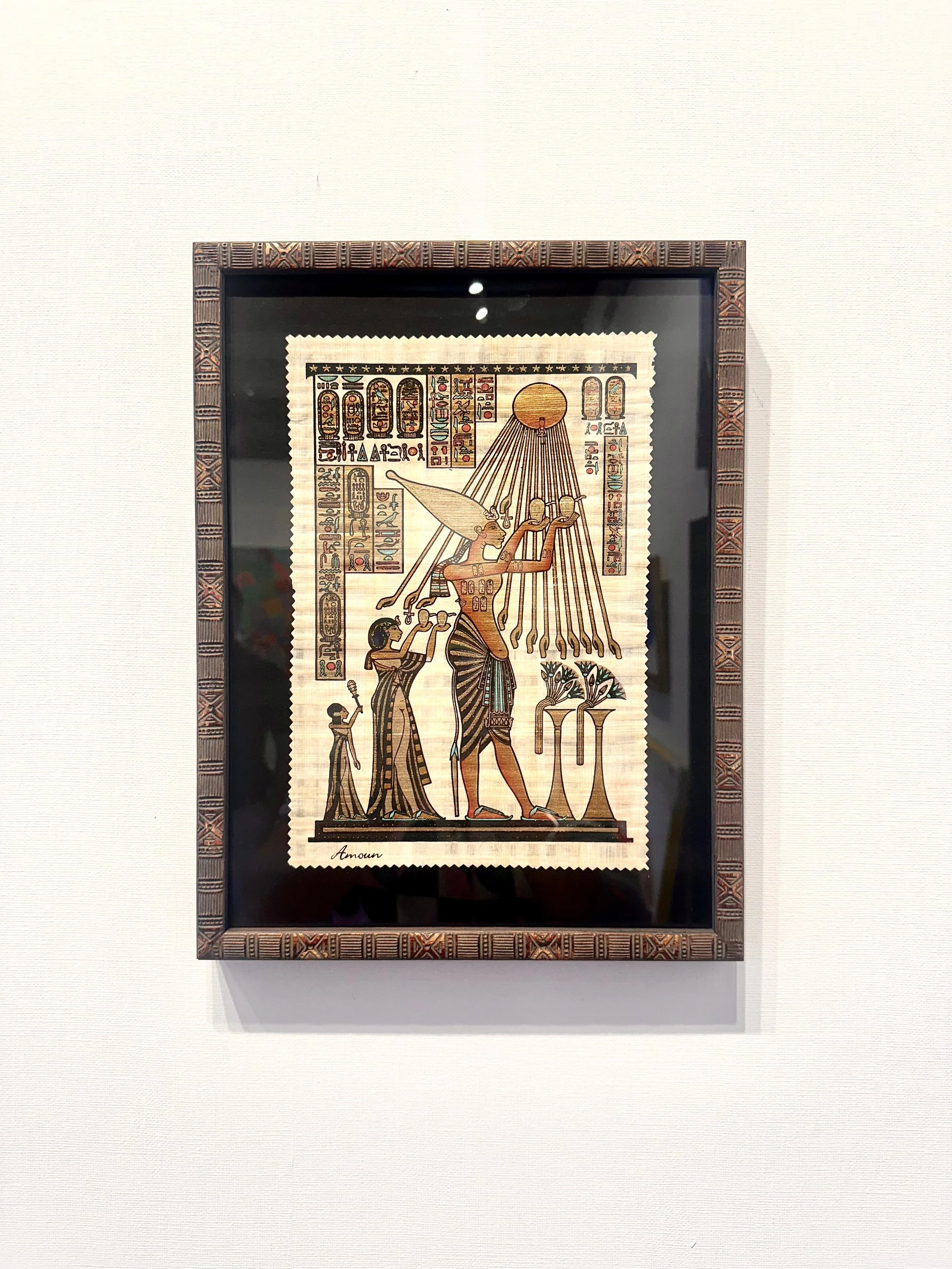 Framed Egyptian-style artwork depicting a god with a sun disk, rays, and a headdress, with smaller figures and hieroglyphs.