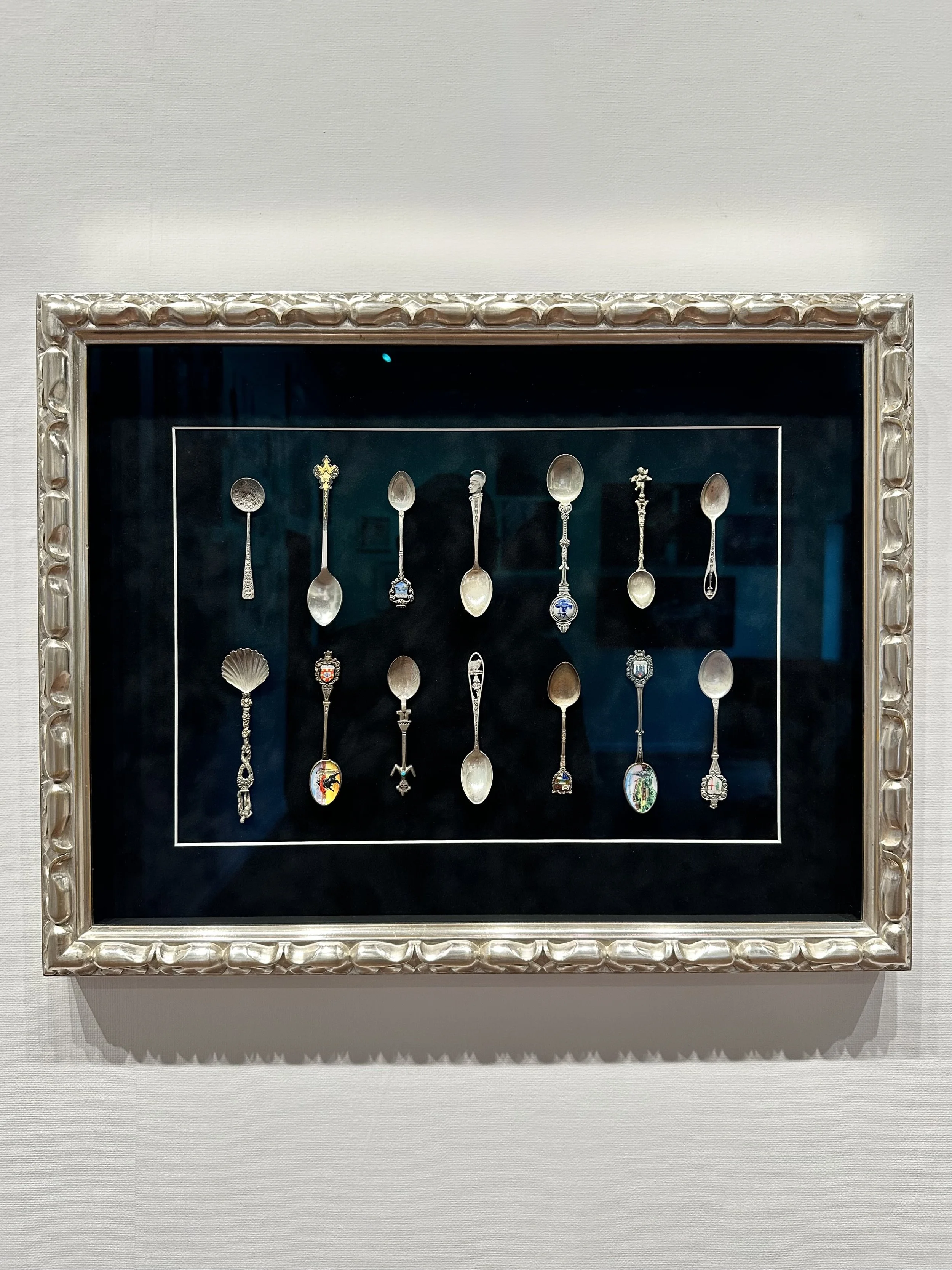 Display of decorative souvenir spoons with various handles and designs in a framed case.