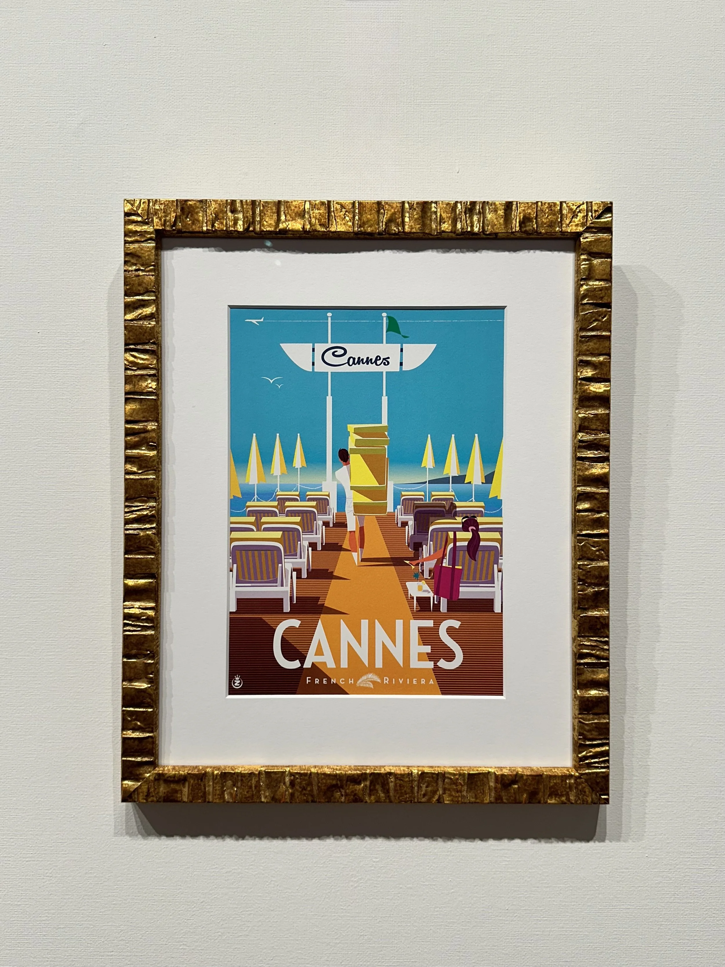 Framed travel poster for Cannes, featuring a beach scene with chairs, umbrellas, and two women in the foreground, with a clear blue sky and seagulls in the background.