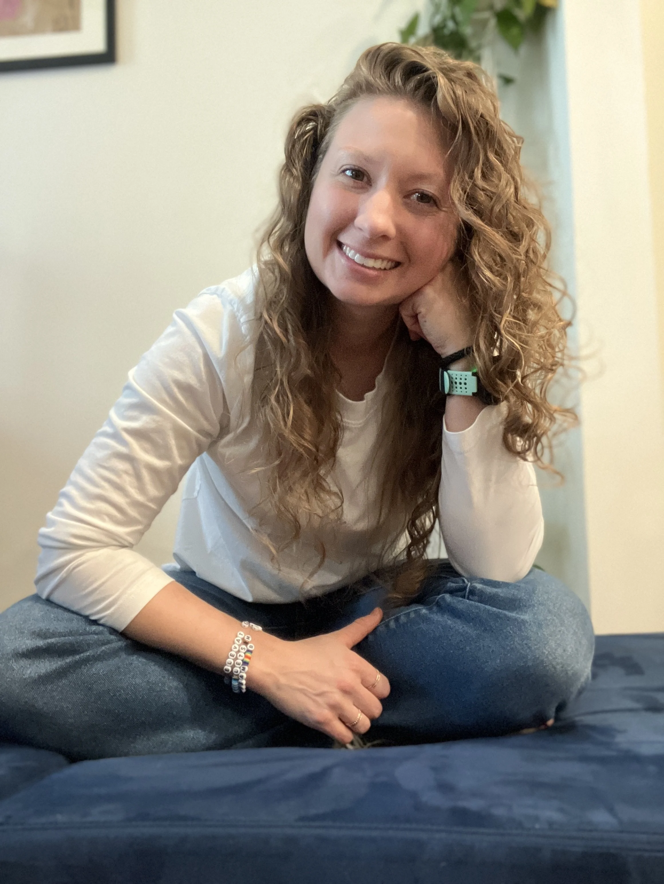 A woman with curly hair sitting on a bed, smiling with her head resting on her hand, wearing a white long-sleeved shirt, jeans, and a bracelet.