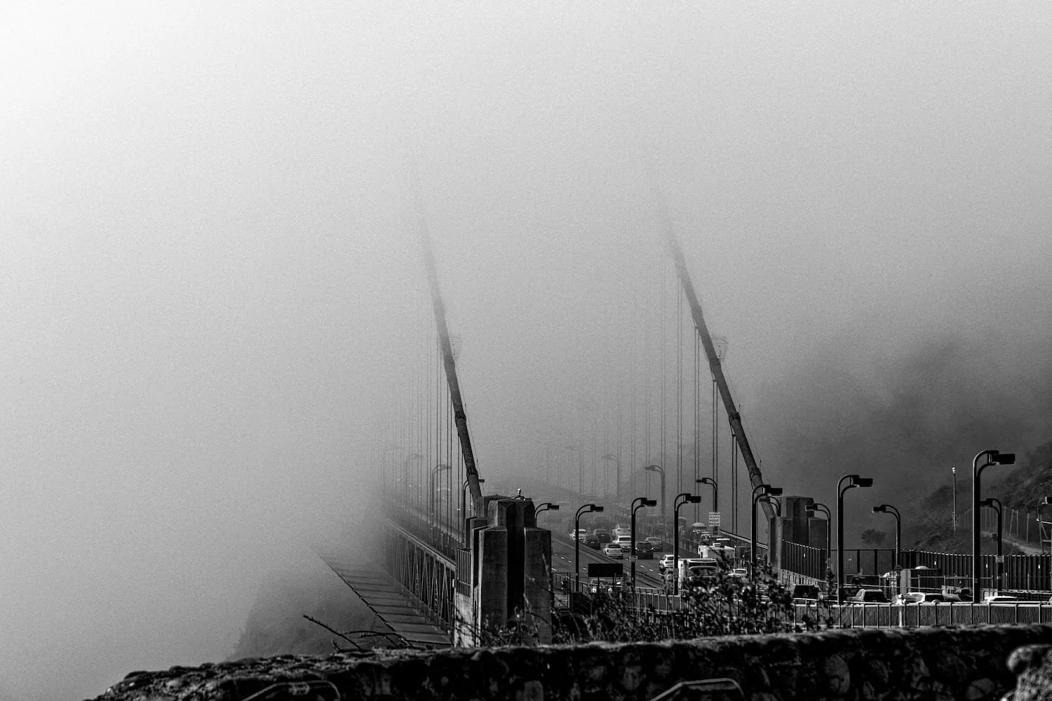 Black and white photo of a suspension bridge shrouded in dense fog with traffic and street lamps visible on the bridge.