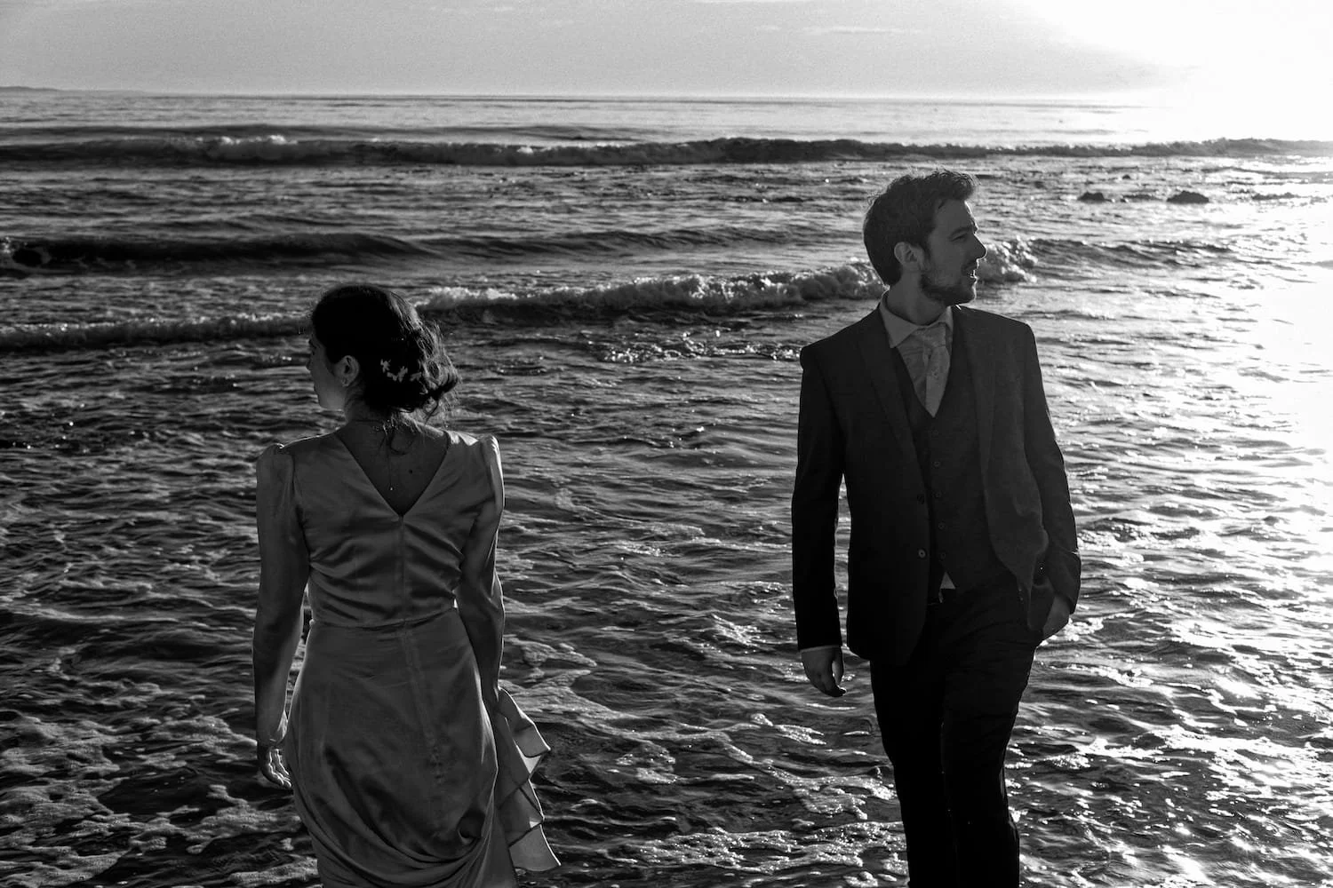 A man and woman in formal attire walking along the shoreline at sunset or sunrise, with ocean waves behind them.