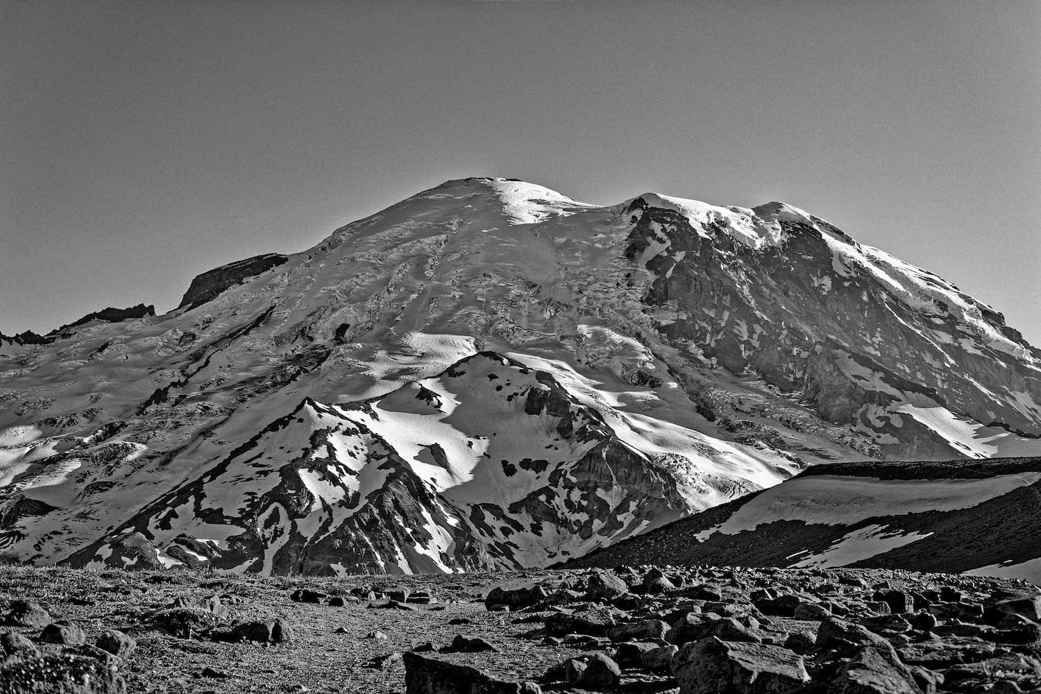 Black and white photo of a snow-covered mountain with rocky terrain in the foreground and clear sky.
