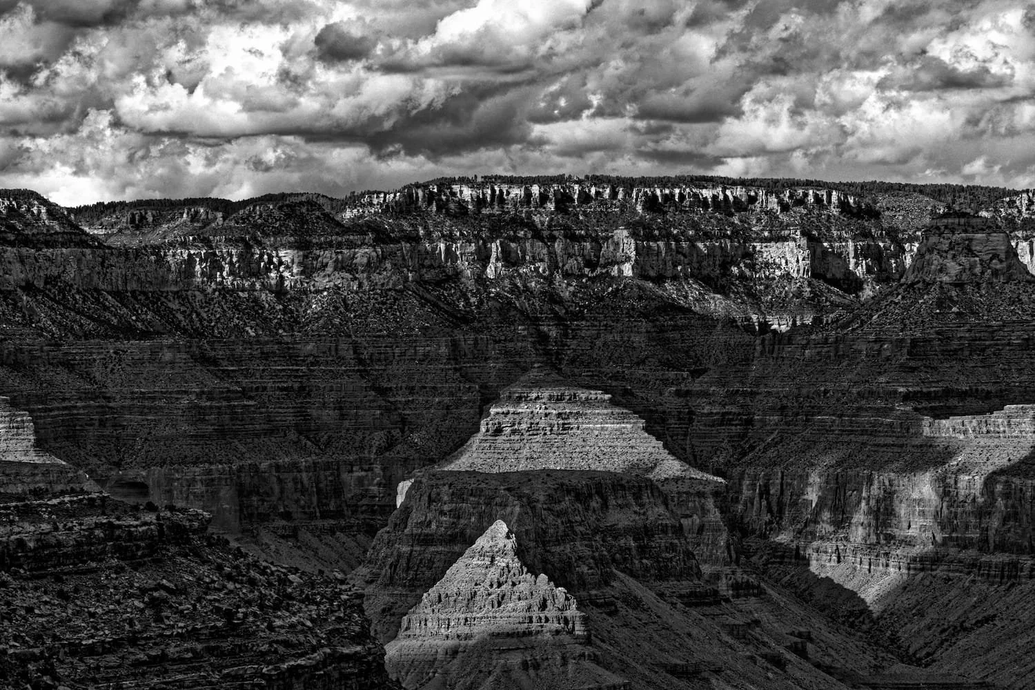 Black and white photograph of the Grand Canyon with layered rock formations, steep cliffs, and cloudy skies above.