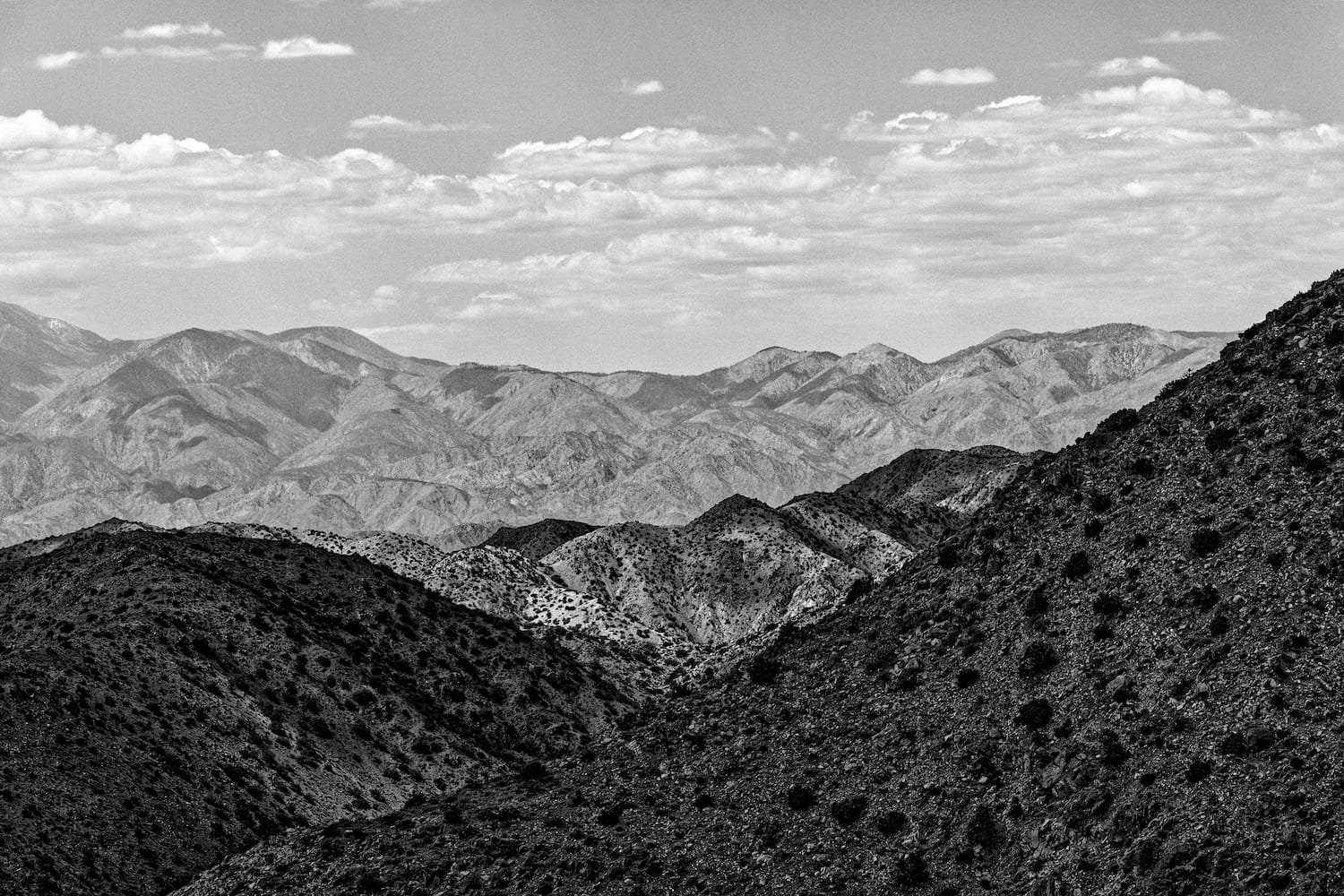 Black and white landscape of mountains with multiple ridges and peaks, some covered in patches of snow, under a sky with scattered clouds.