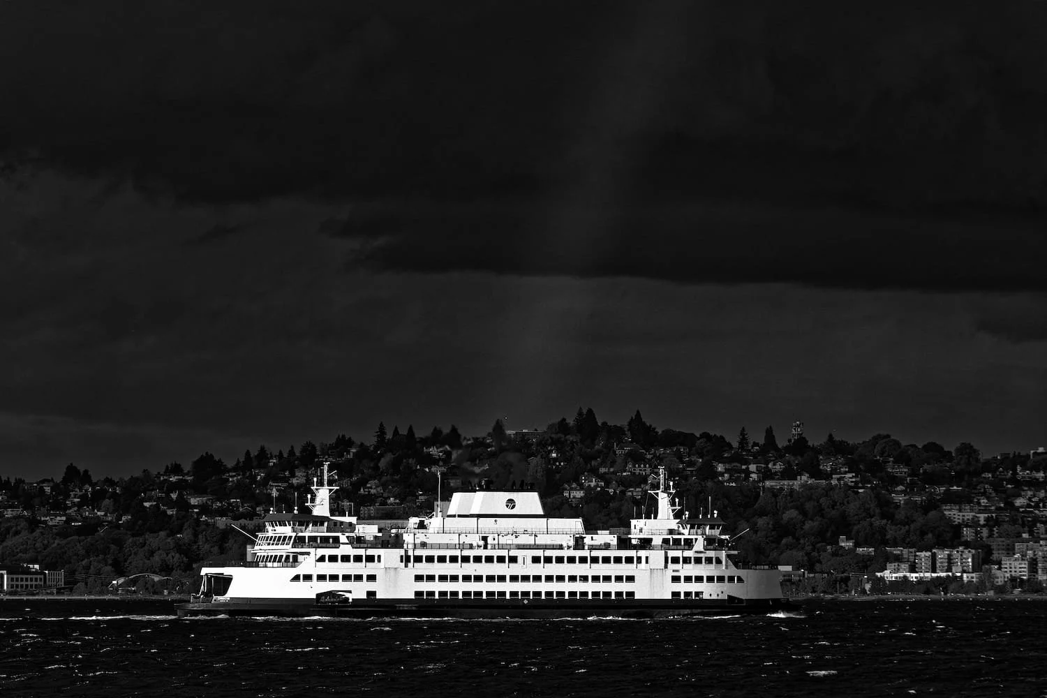 A large white ferry boat sailing on dark water with a cityscape and hillside in the background under a cloudy sky.