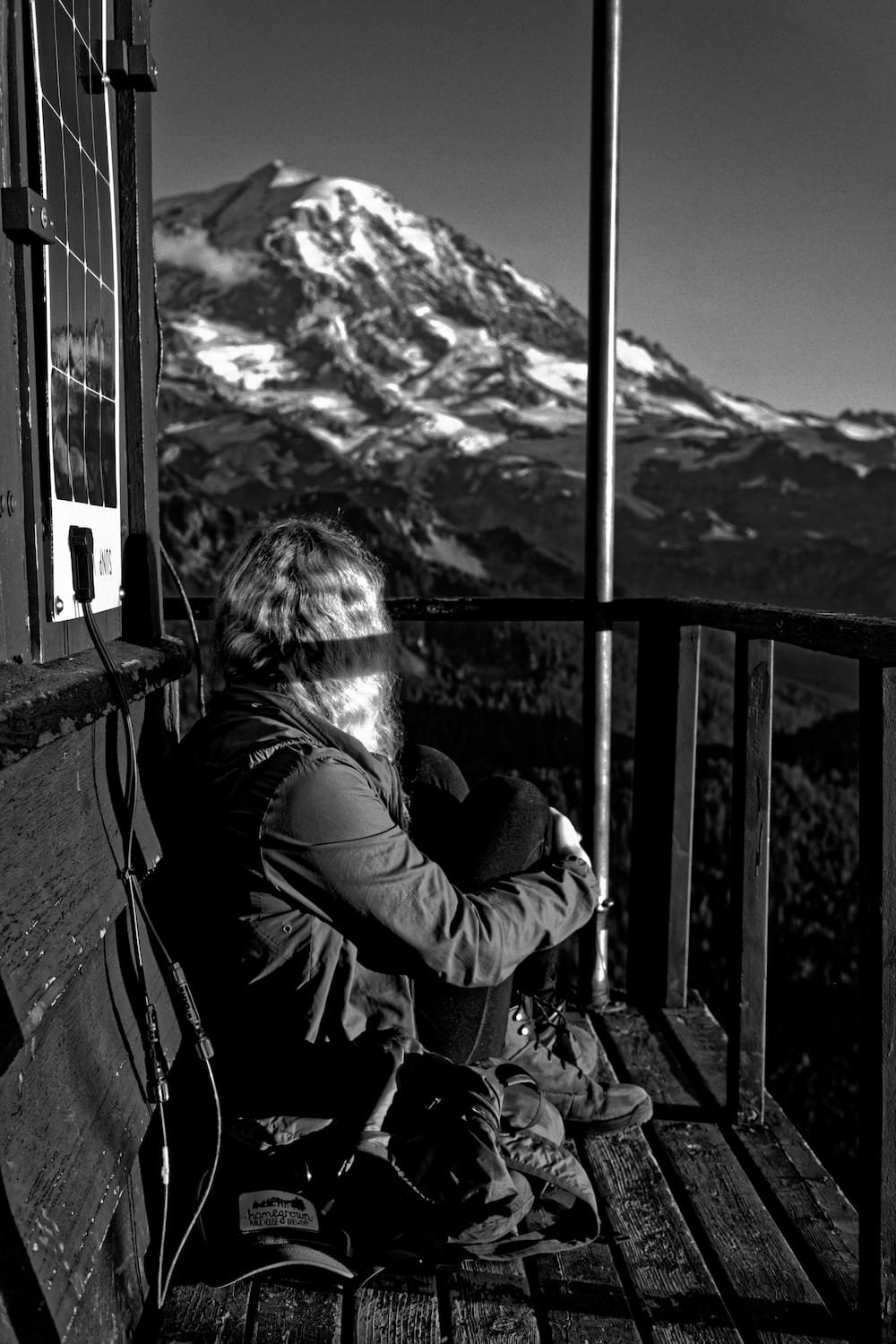 Person sitting on a wooden observation deck, gazing at a snow-capped mountain, with some equipment nearby, in black and white