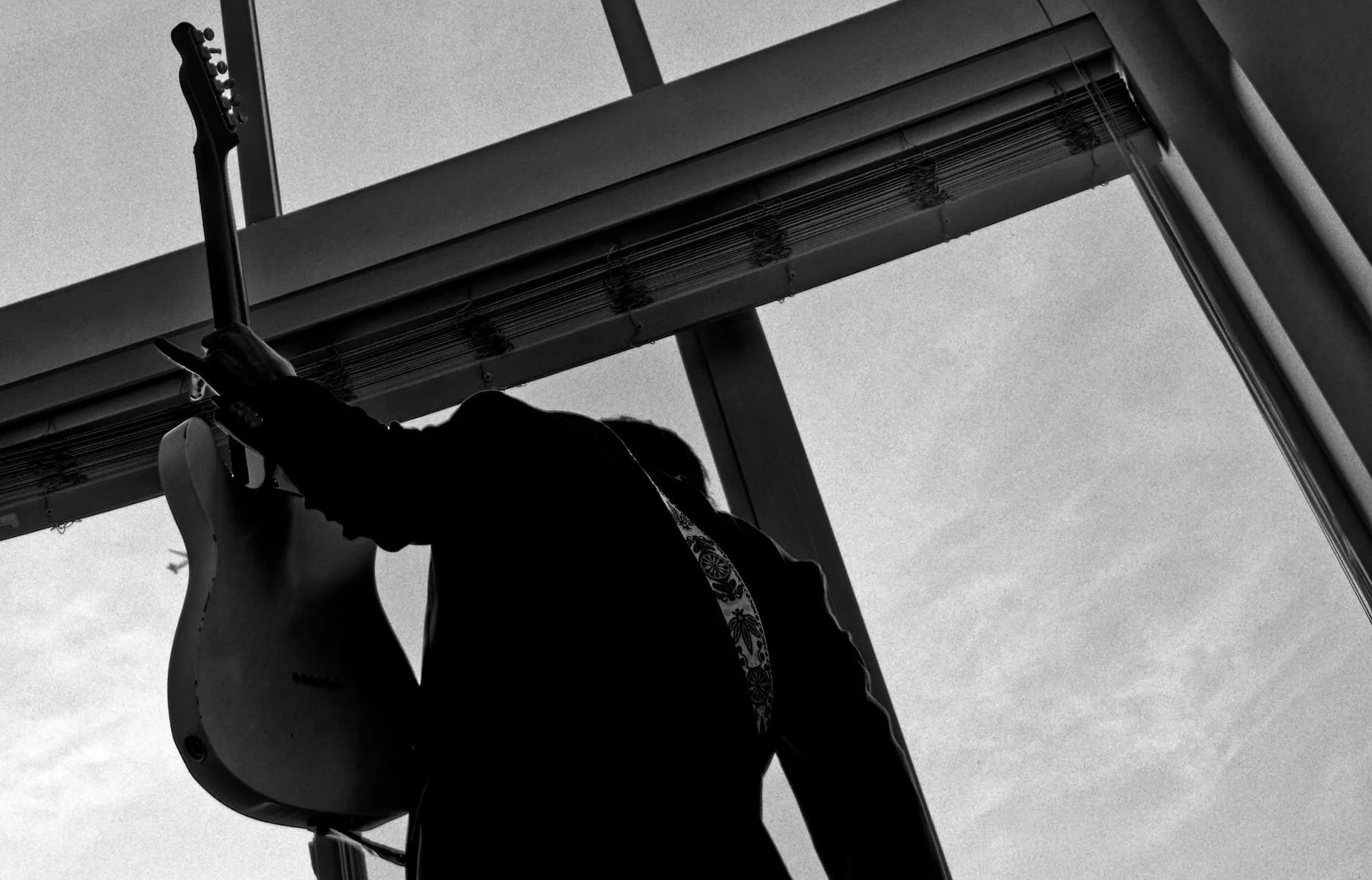 Silhouette of a person holding an electric guitar, standing near a large window, with a view of the sky outside.