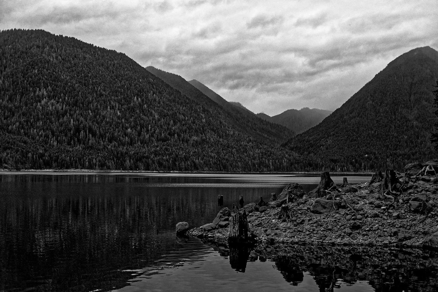A black and white photograph of a mountain landscape with a calm lake in the foreground. The scene features densely forested mountains with a cloudy sky above.
