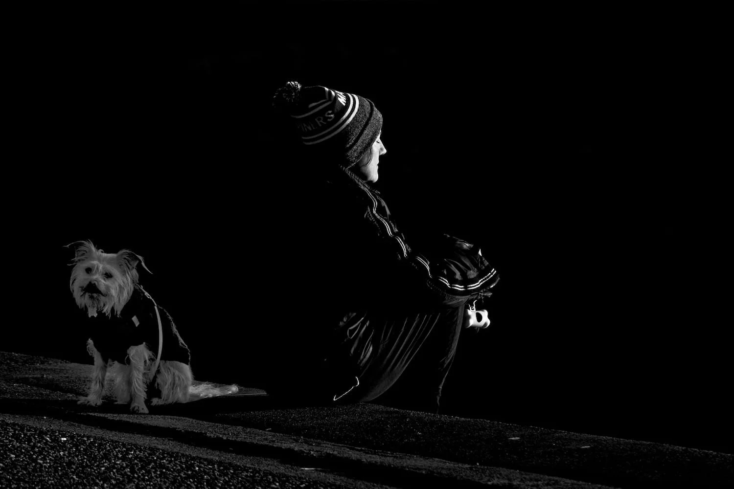 A person sitting outdoors at night with a dog. The person is wearing a winter hat and jacket, facing sideways, with arms crossed. The dog is sitting on the ground and looking towards the camera.