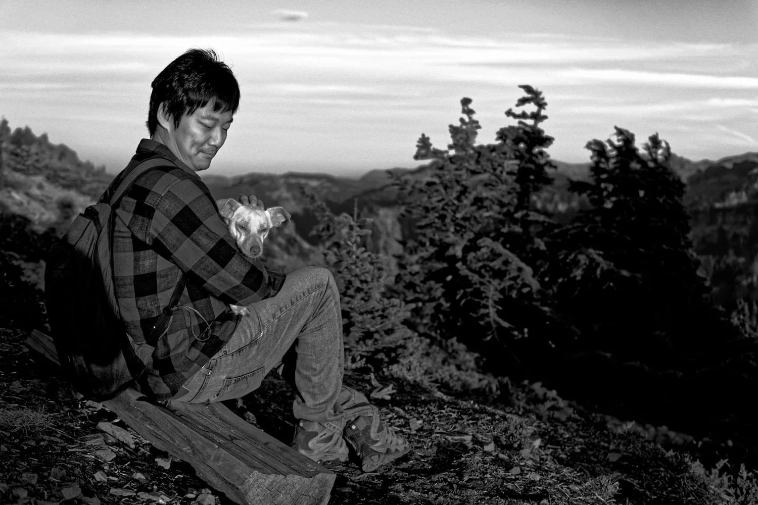 A man in a plaid shirt sitting on a log holding a dog in a forested area with mountains in the background