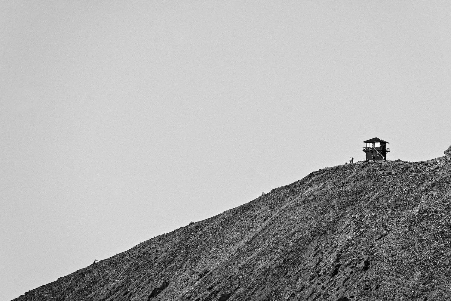 A black and white photo of a hill or mountain with a small watchtower and two people standing near it at the top.