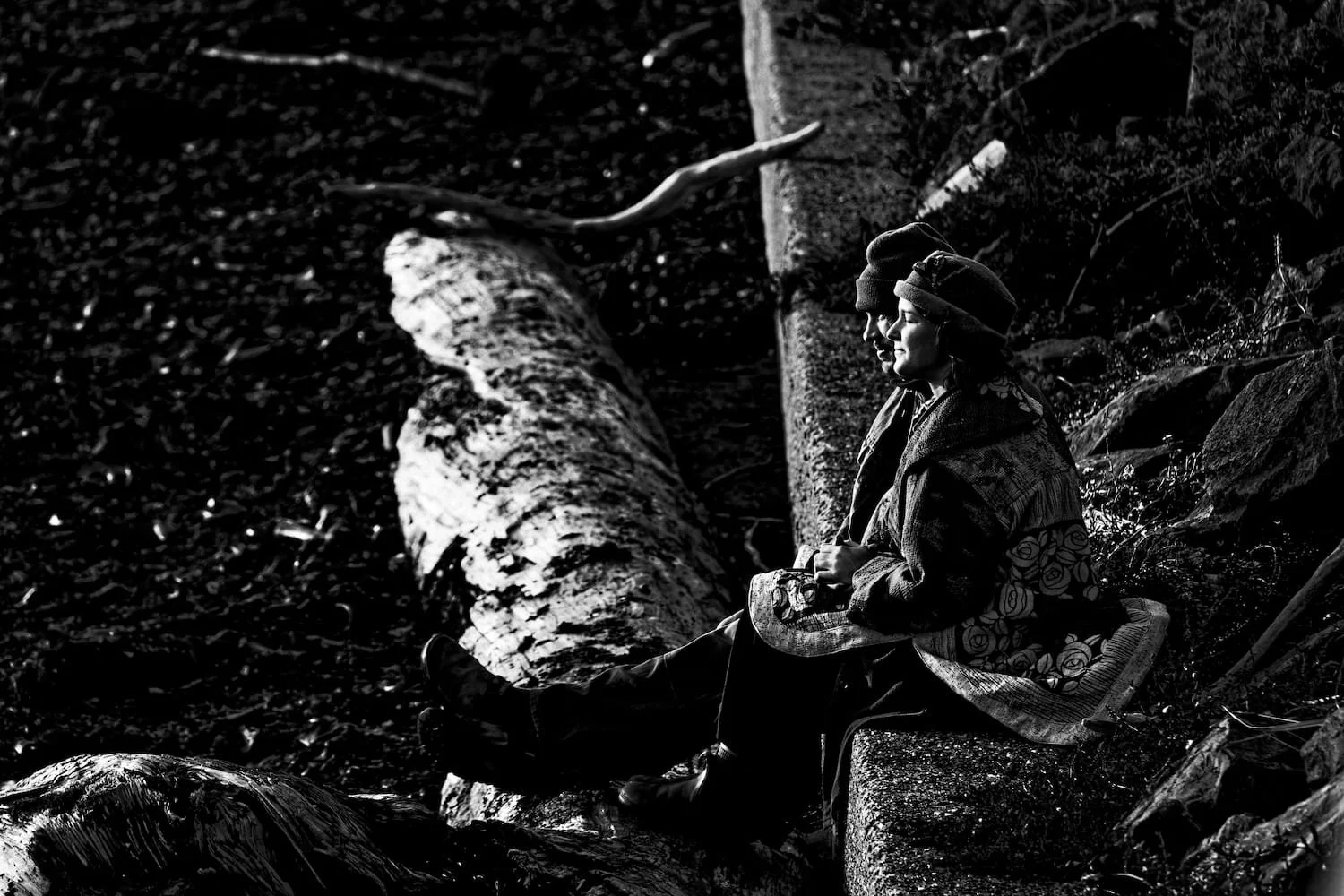 A black and white photo of two children sitting on a rock by a stream, wearing winter clothing and beanies, with a fallen tree in the background.