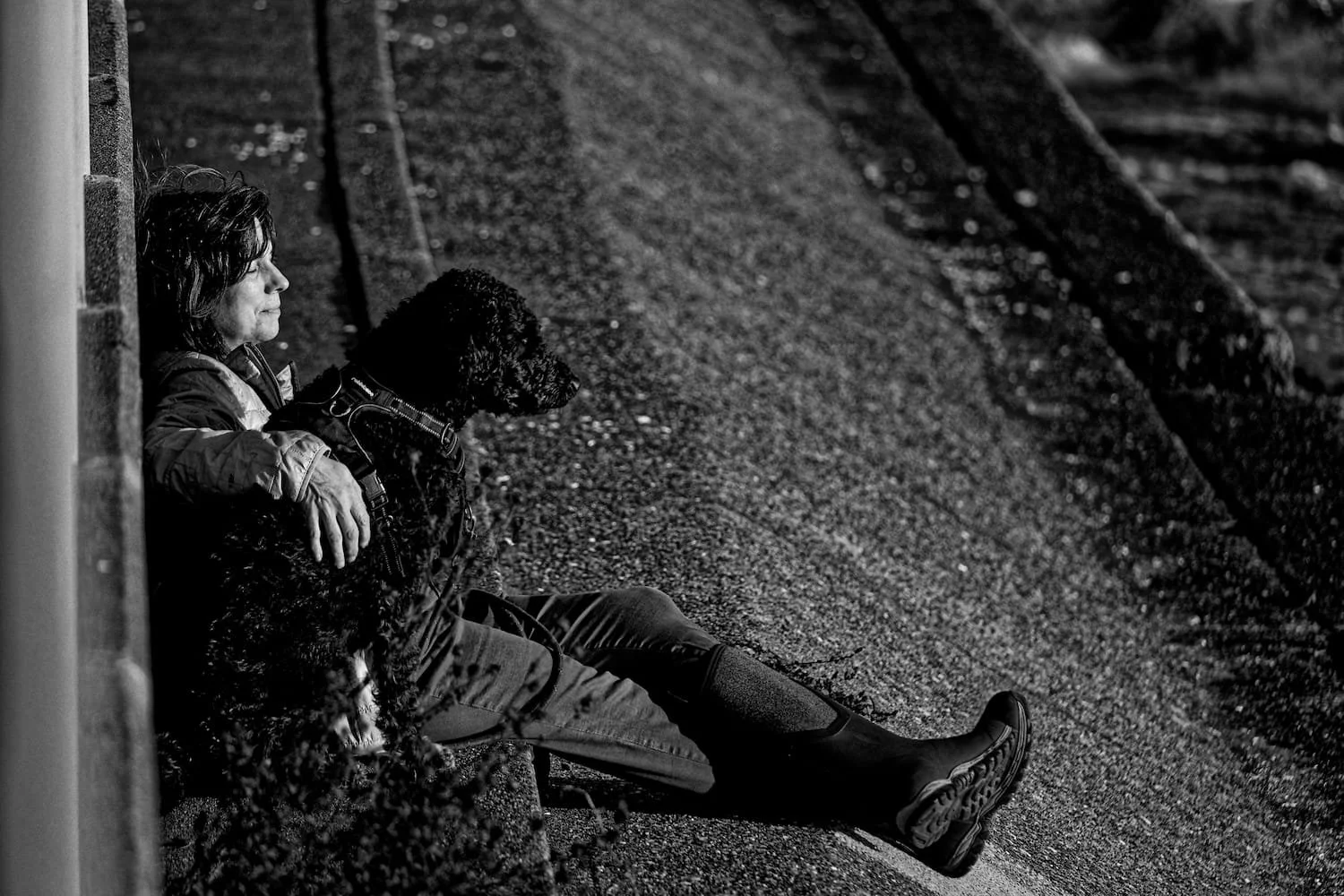 A woman and her dog sitting on the sidewalk, both looking in the same direction, with the woman leaning against a wall and the dog between her and the sidewalk.