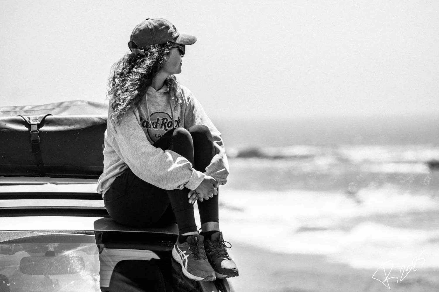 A woman sitting on the back of a car, looking towards the beach and ocean in the distance. She is wearing a cap, sunglasses, a hoodie, and sneakers, with a bag beside her.