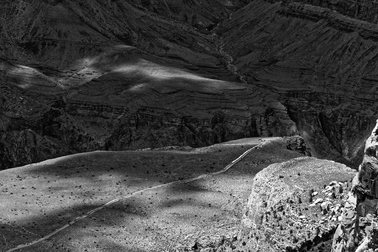 A black and white photo of a winding trail through a barren, rocky canyon landscape with steep cliffs and layered geological formations.