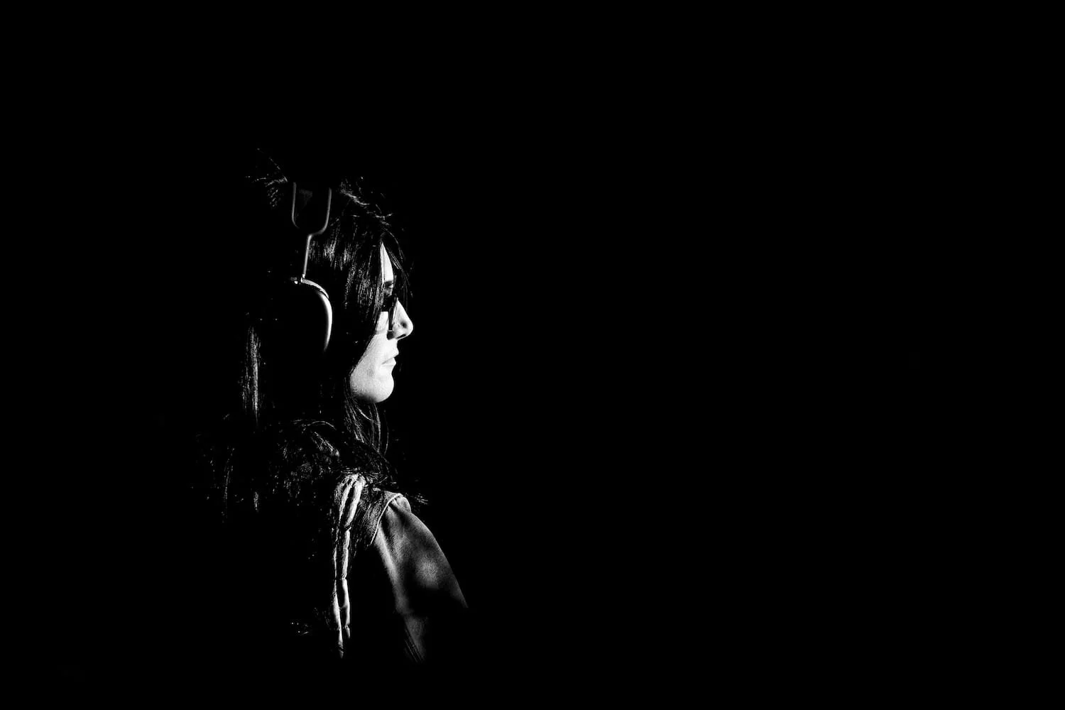 Side profile of a young woman with glasses, long dark hair, and a leather jacket, wearing headphones, against a black background.