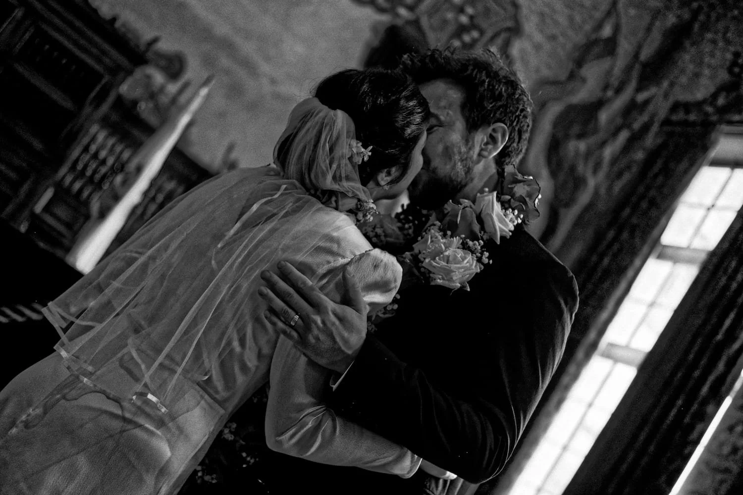 A black-and-white photo of a bride and groom kissing during their wedding, both wearing floral arrangements around their necks, inside a rustic house with a window in the background.